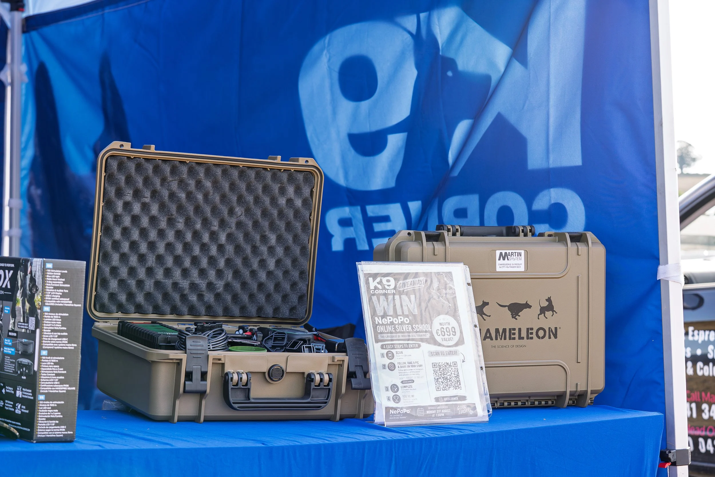 A display table with equipment and signage for a giveaway event, featuring a tan plastic case with electronic devices inside and a sign indicating a chance to win $699, at an outdoor event with a blue backdrop.