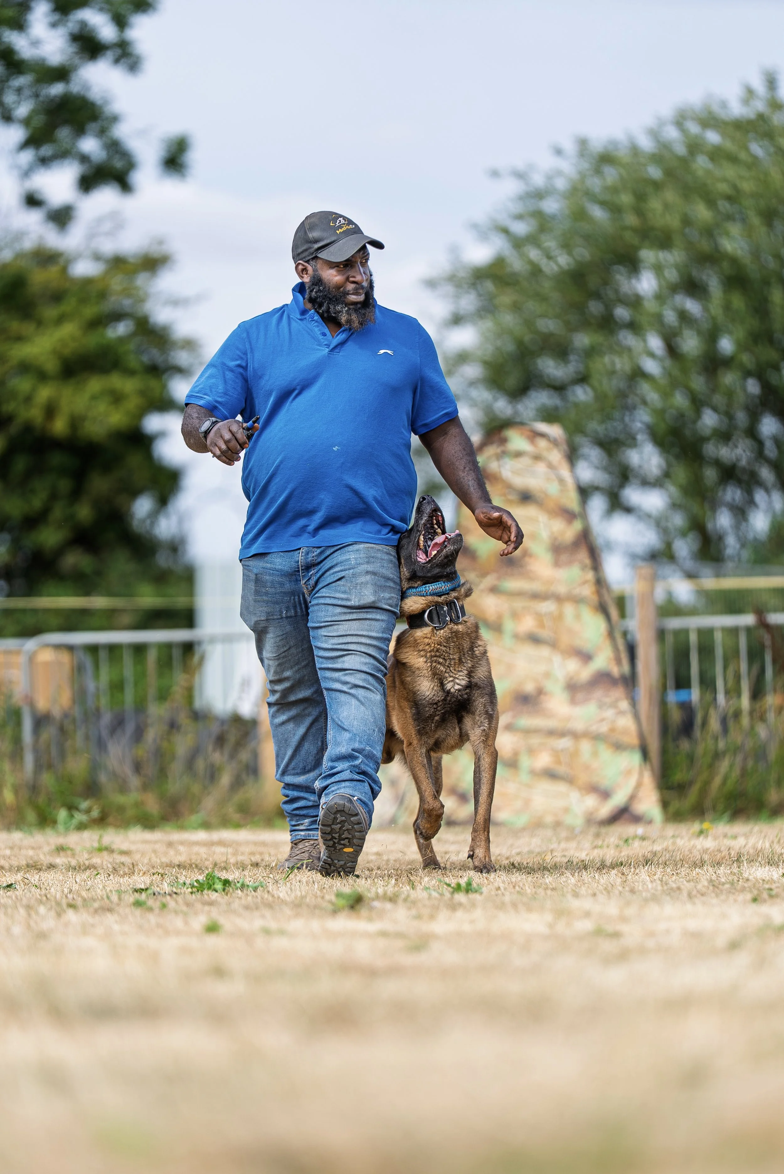 A man in a blue shirt and cap walking a German Shepherd on a track outdoors.
