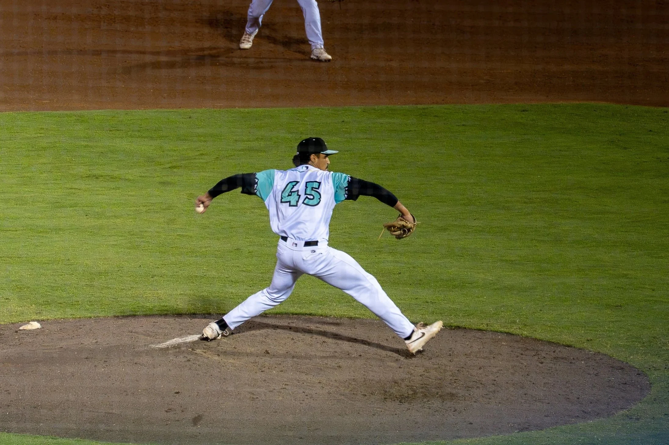 A baseball player in a white uniform with the number 45 on his back is fielding a ground ball near the pitcher's mound, with another player visible in the background.