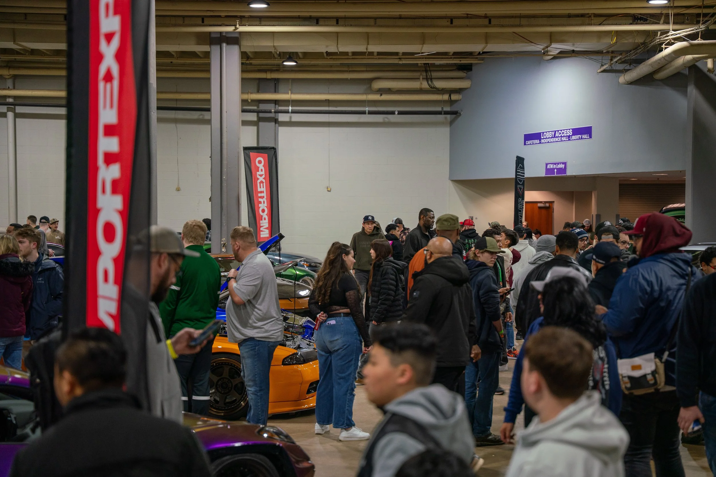 Crowded indoor automotive eventwith attendees viewing custom cars and displays.