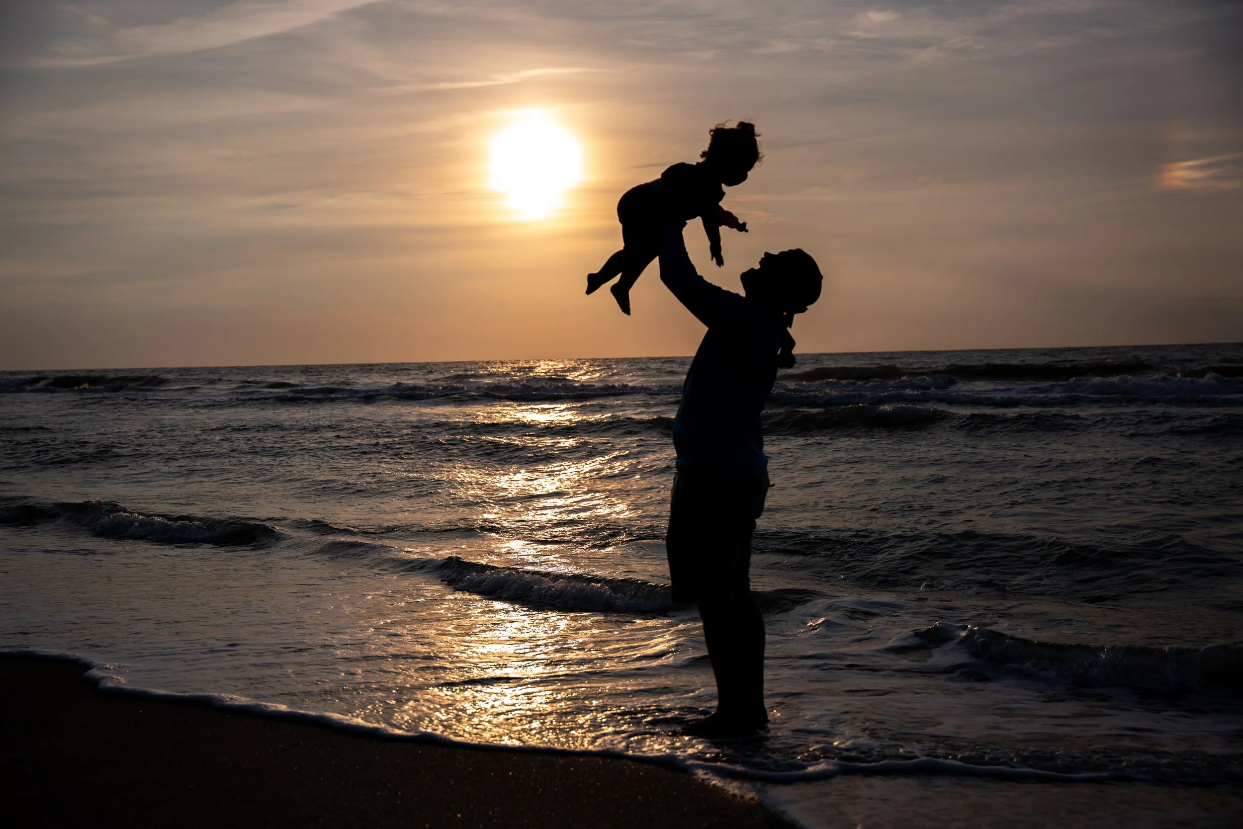 Silhouette of an adult lifting a child into the air on the beach during a golden sunset. 