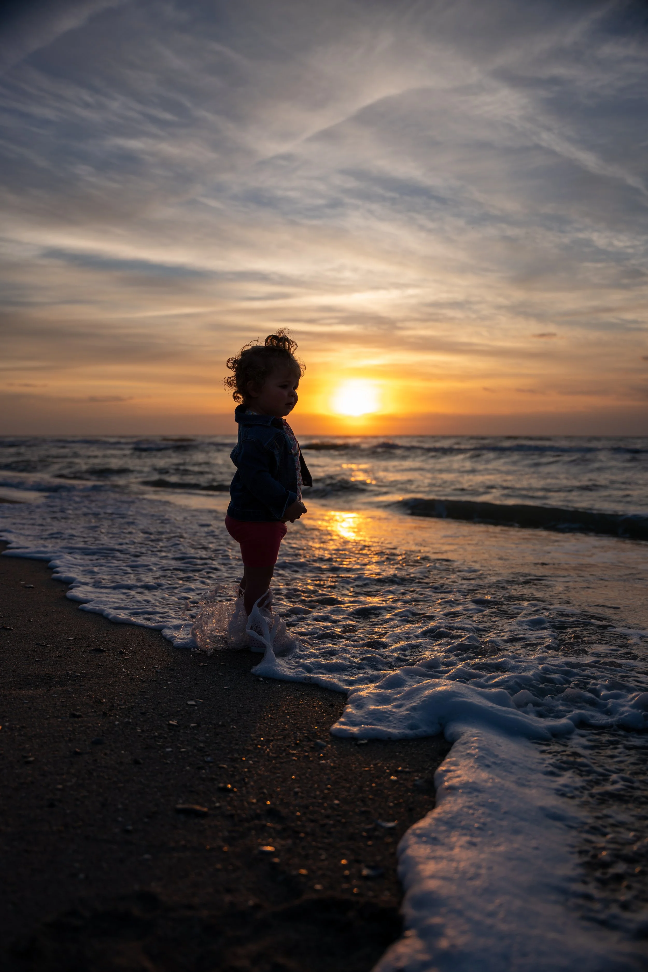Young girl standing along the shoreline at sunset with ocean waves in the background. 