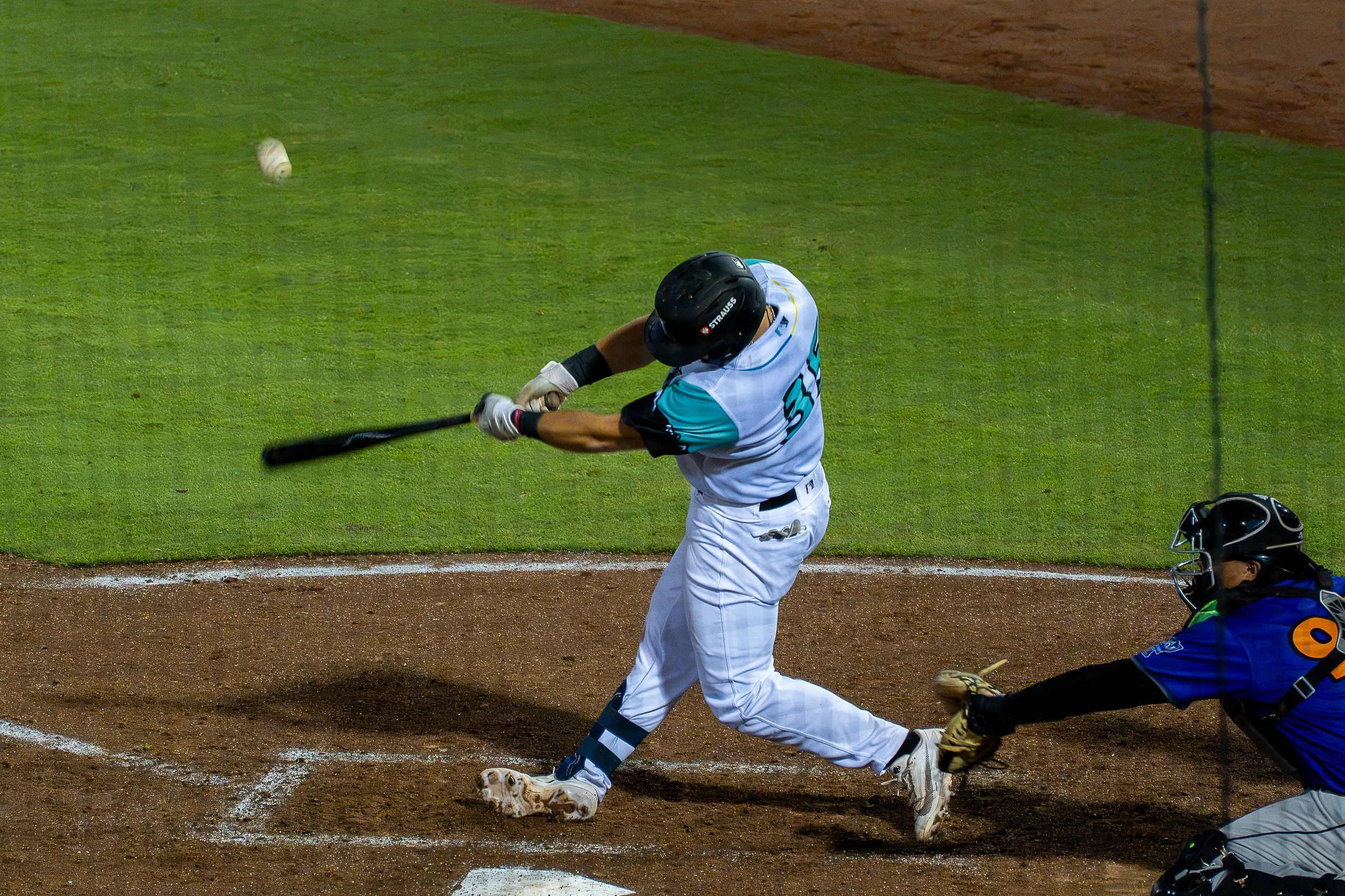 A baseball player in a white and teal uniform swinging a bat at a pitched ball near home plate during a game.