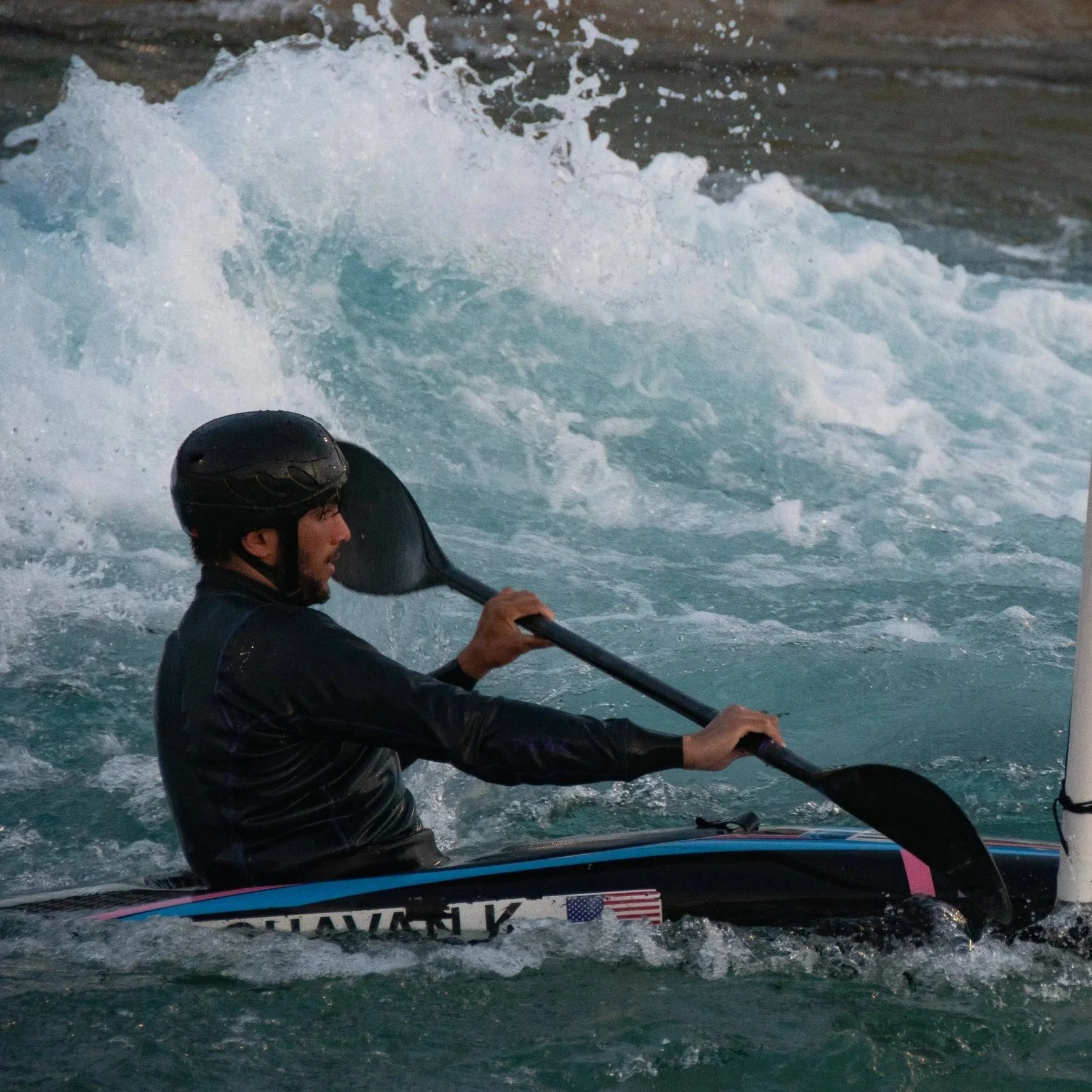 Man wearing a helmet kayaking in turbulent water with white splashes and foam.