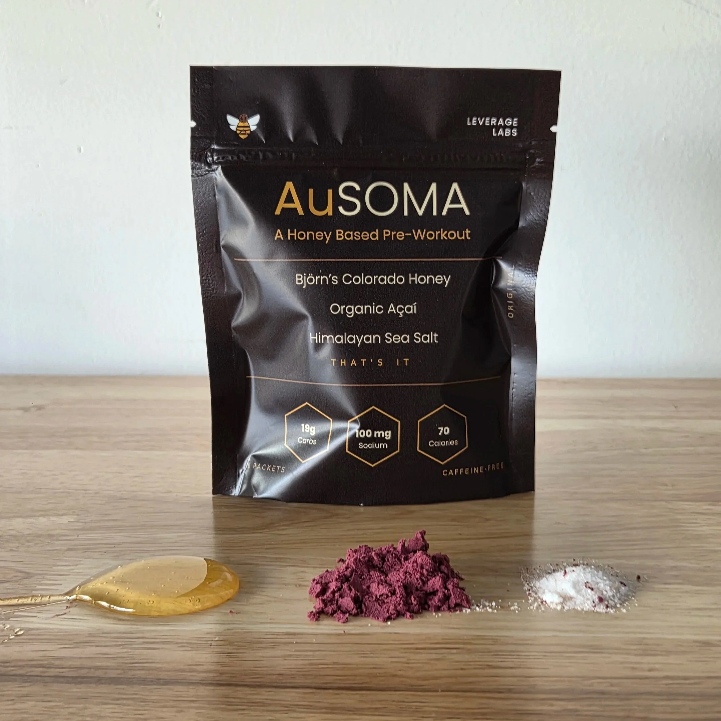 A black and gold package of Ausoma honey-based pre-workout powder on a wooden surface with three piles of powders in yellow, purple, and white colors in front of it.