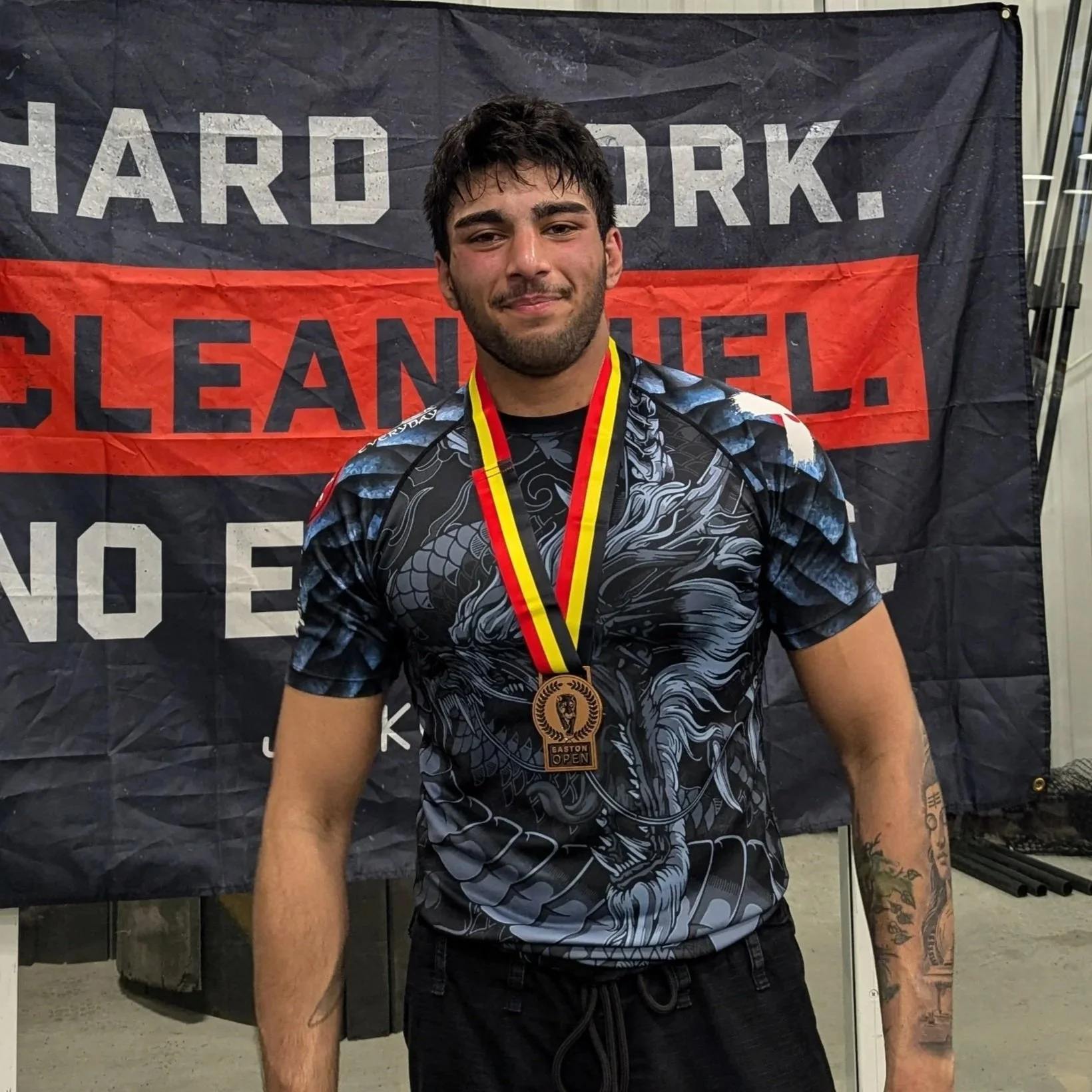 A young man with dark hair and a beard, smiling, wearing a black and gray patterned sports shirt and black shorts, with a gold medal around his neck, standing in front of a black and red banner that reads 'HARD WORK. CLEANLINESS.' on a gray concrete floor.