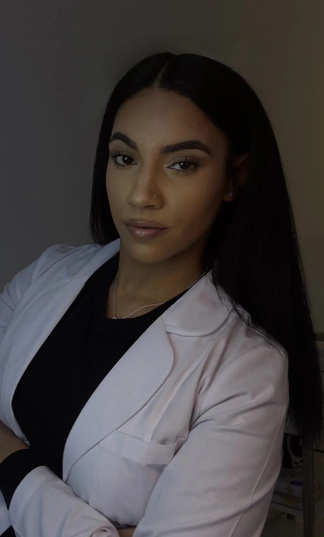 A woman with long dark hair, light brown skin, wearing a white lab coat over a black top, posing with arms crossed in a dimly lit environment.