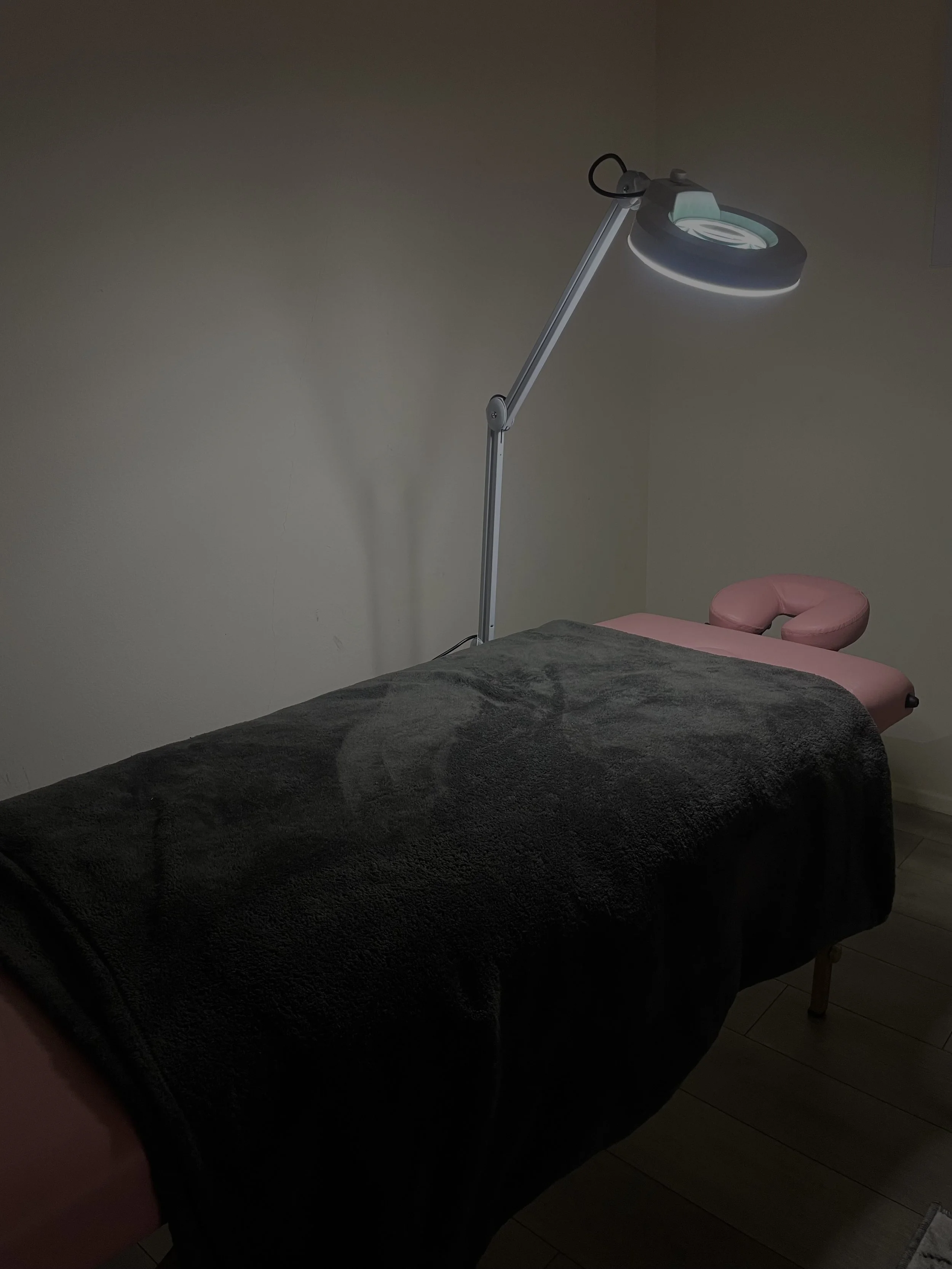 A massage or therapy room with a bed covered by a dark blanket, a pink massage or therapy chair at the head of the bed, an adjustable overhead light, and a plain wall.
