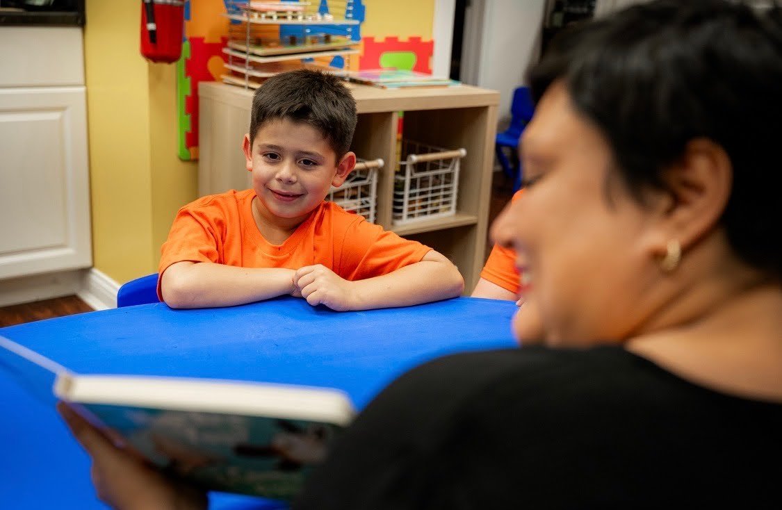 A young boy with short dark hair in an orange shirt sitting at a blue table, smiling and looking at an adult woman reading a book, with a classroom setting in the background.
