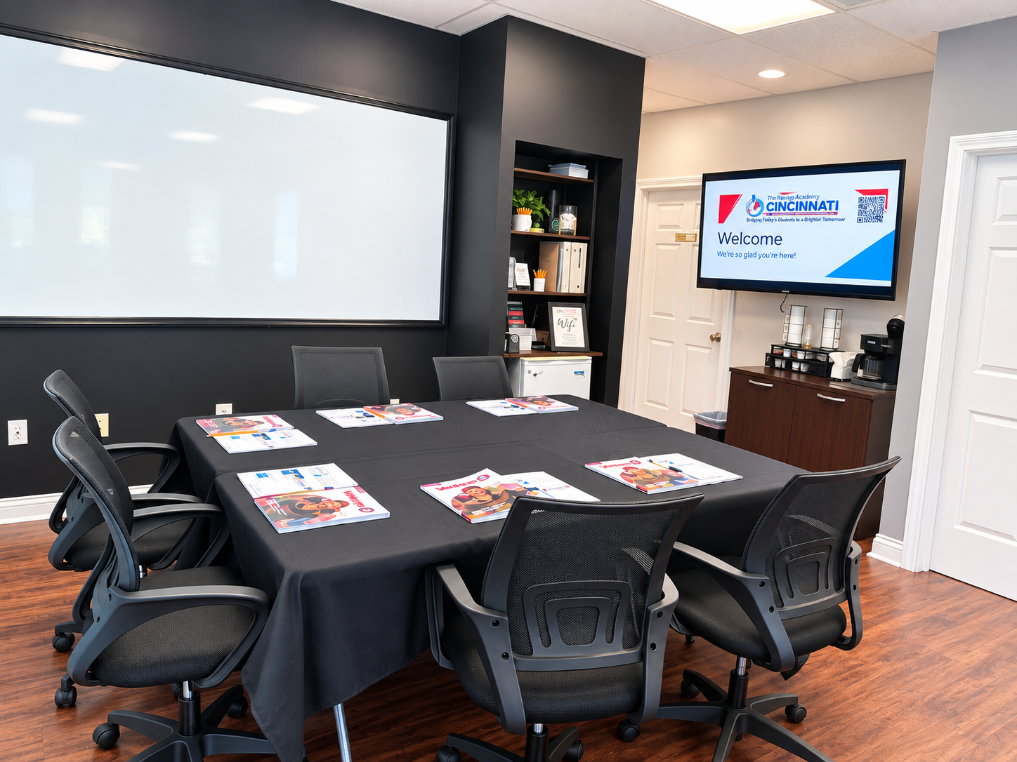 Meeting room with a rectangular table covered with a black tablecloth, six chairs around it, and brochures on the table. There is a large whiteboard on the wall, a shelf with books and decor, and a wall-mounted screen displaying the Cincinnati Academy logo and welcome message. Coffee accessories are on a cabinet in the corner.