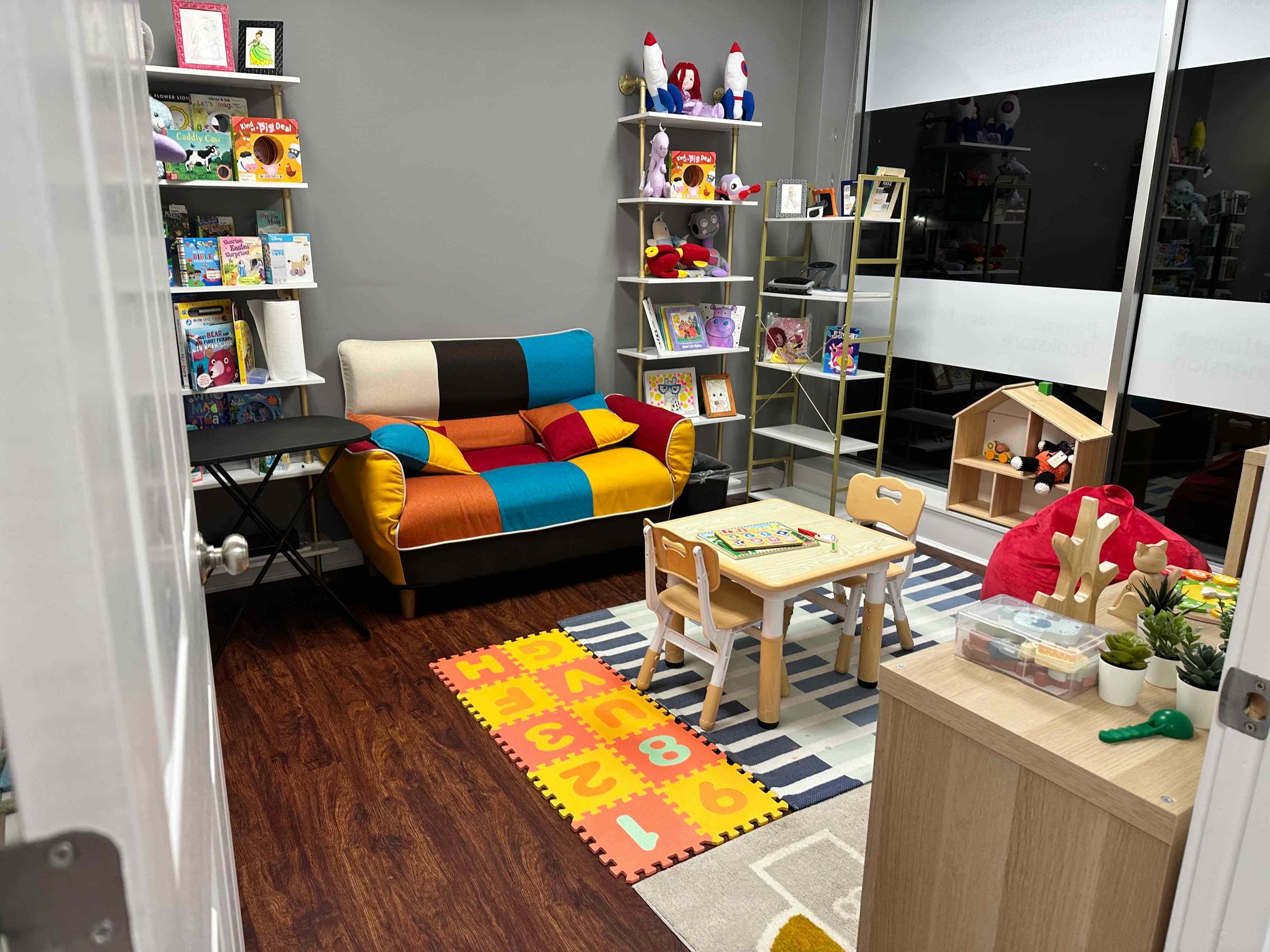 Colorful children's playroom with bookshelves, plush toys, artwork, a small table with chairs, and a foamed alphabet mat on wood flooring.