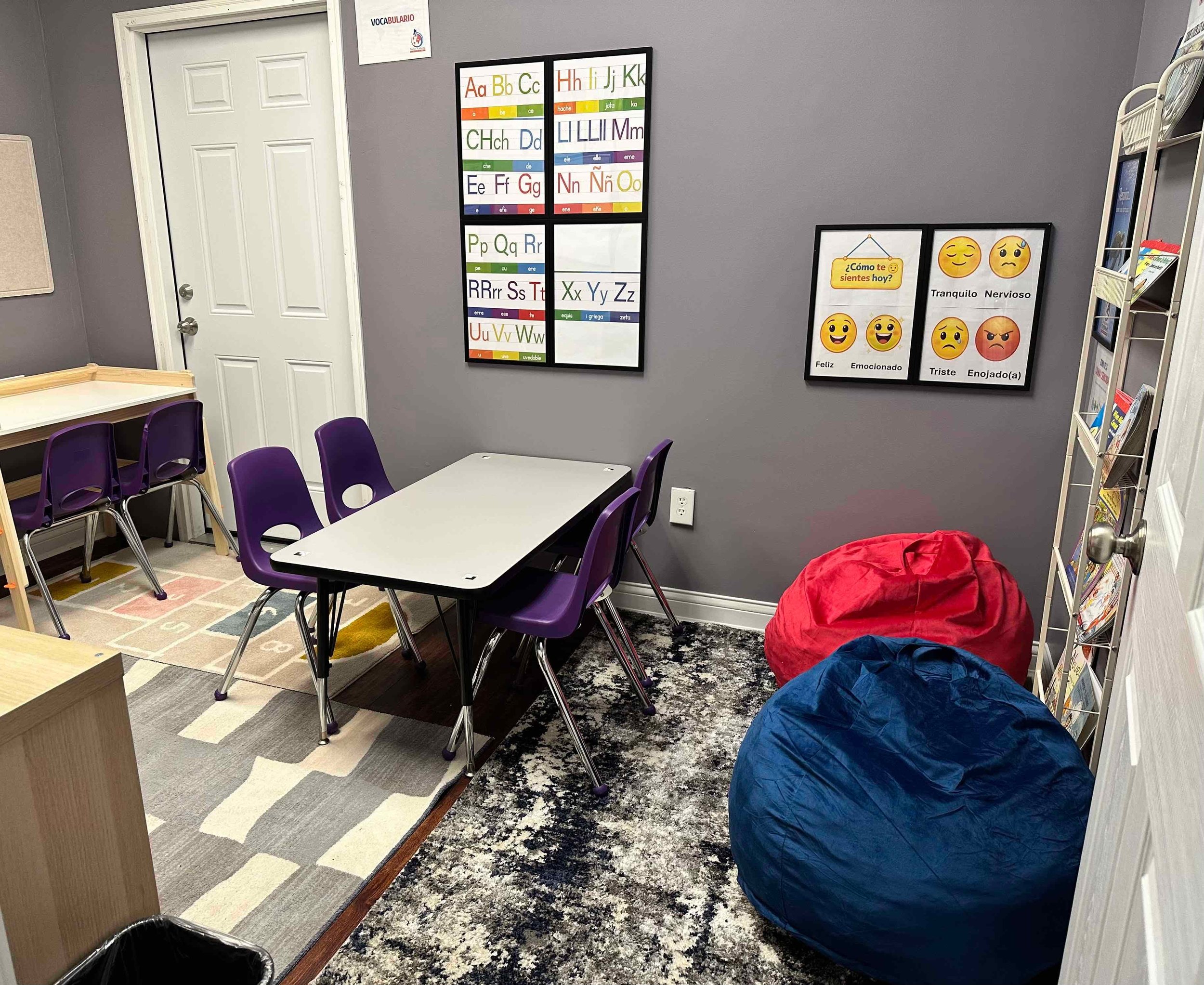 Inside a classroom with purple chairs around white tables, posters on the gray wall with Spanish alphabet and emotion emojis, and colorful rugs, some jackets on the floor near a white shelf holding children's books.