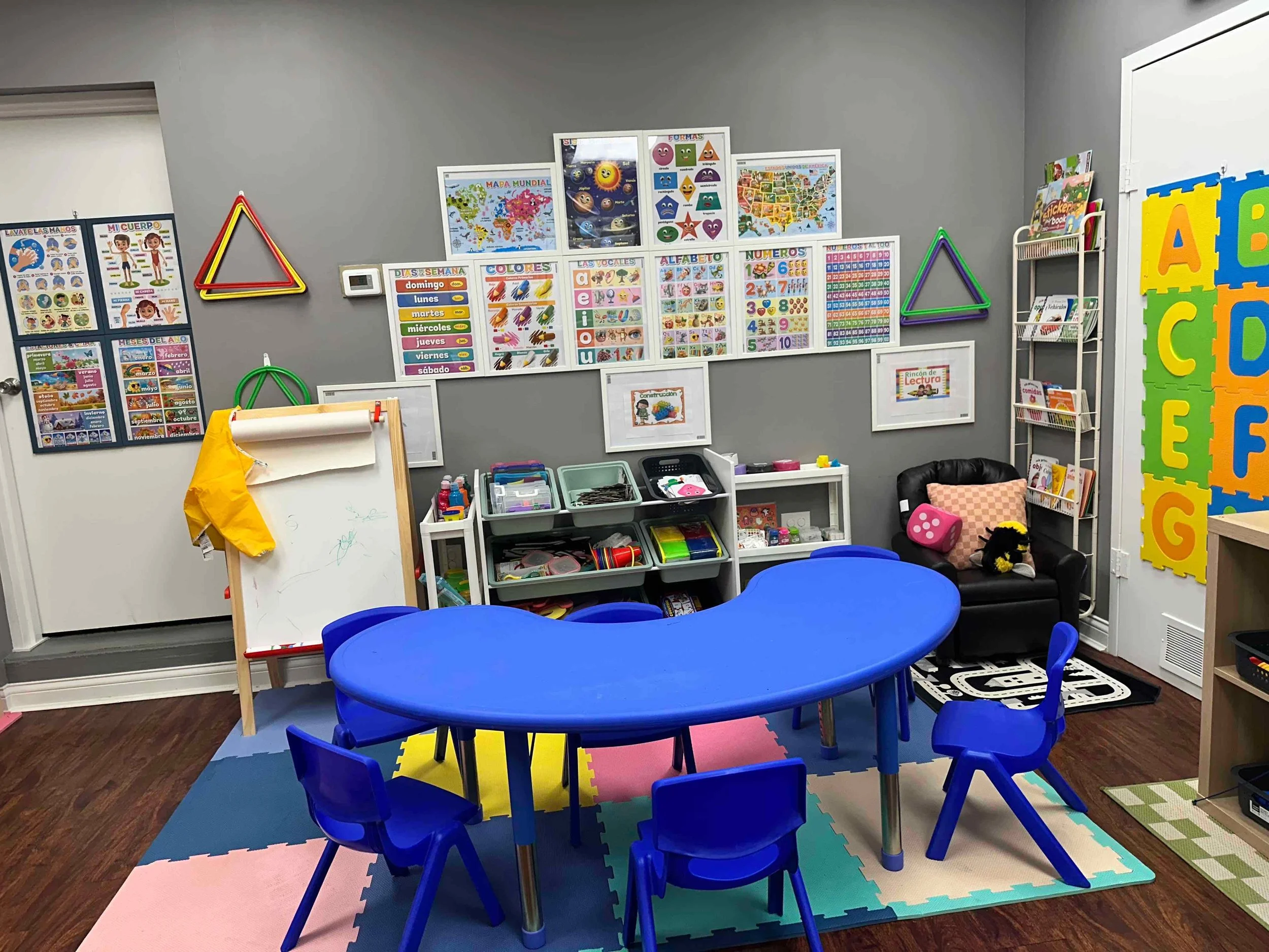 A colorful preschool classroom with a blue table and chairs, educational posters on the walls, a whiteboard, and various toys and books.