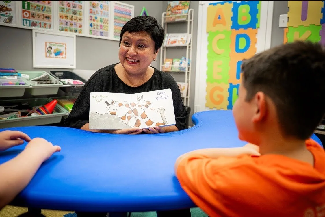 A teacher in a classroom holding a children's book showing two giraffes, while two children sit at a blue table listening. The classroom has colorful alphabet and number posters on the walls.