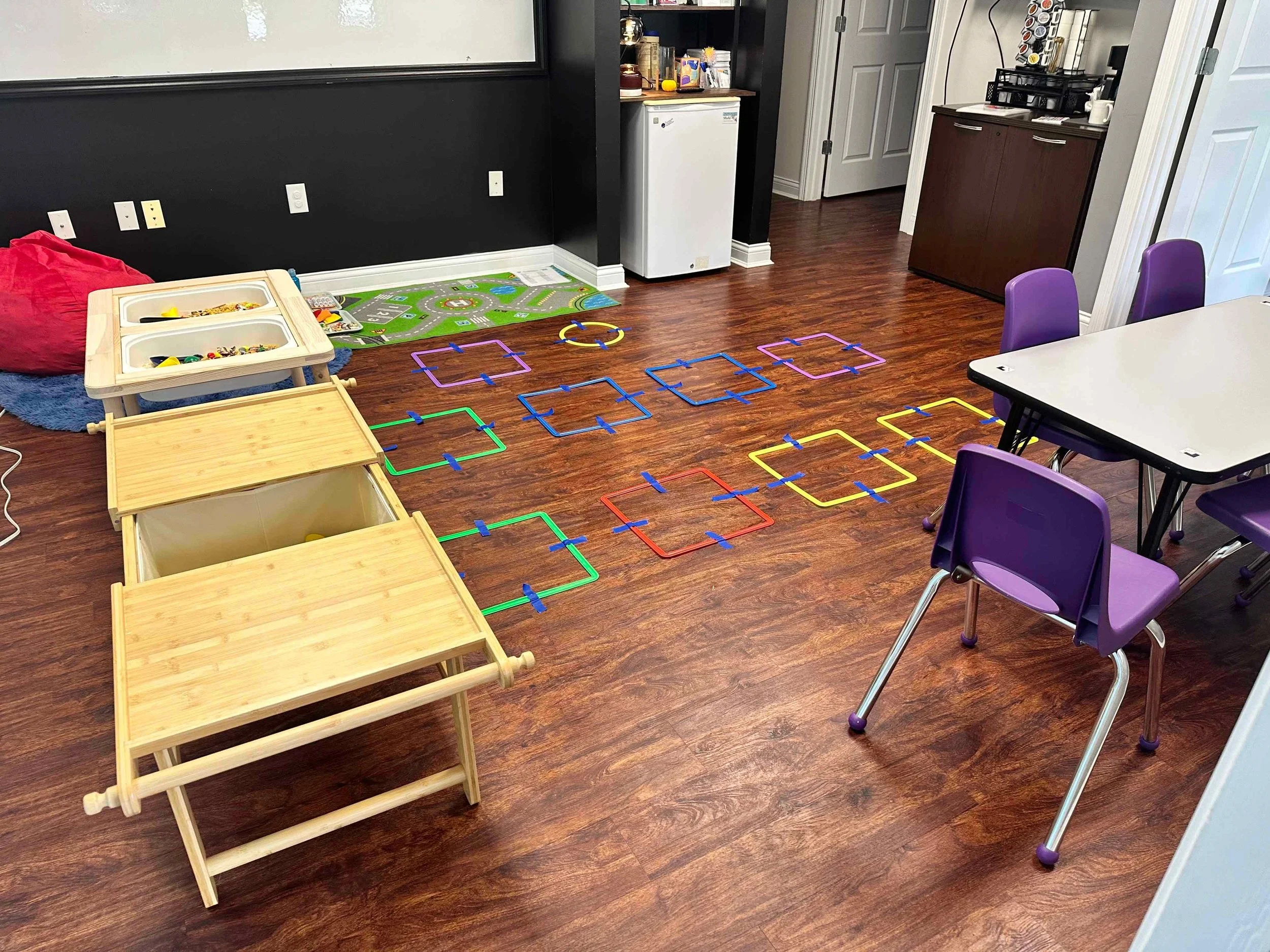A small classroom or playroom with wooden flooring, purple chairs around a white table, a red bean bag, a wooden toy train, and colorful hopscotch squares taped to the floor. There is a black shelf with books and toys, a mini fridge, and a kitchenett