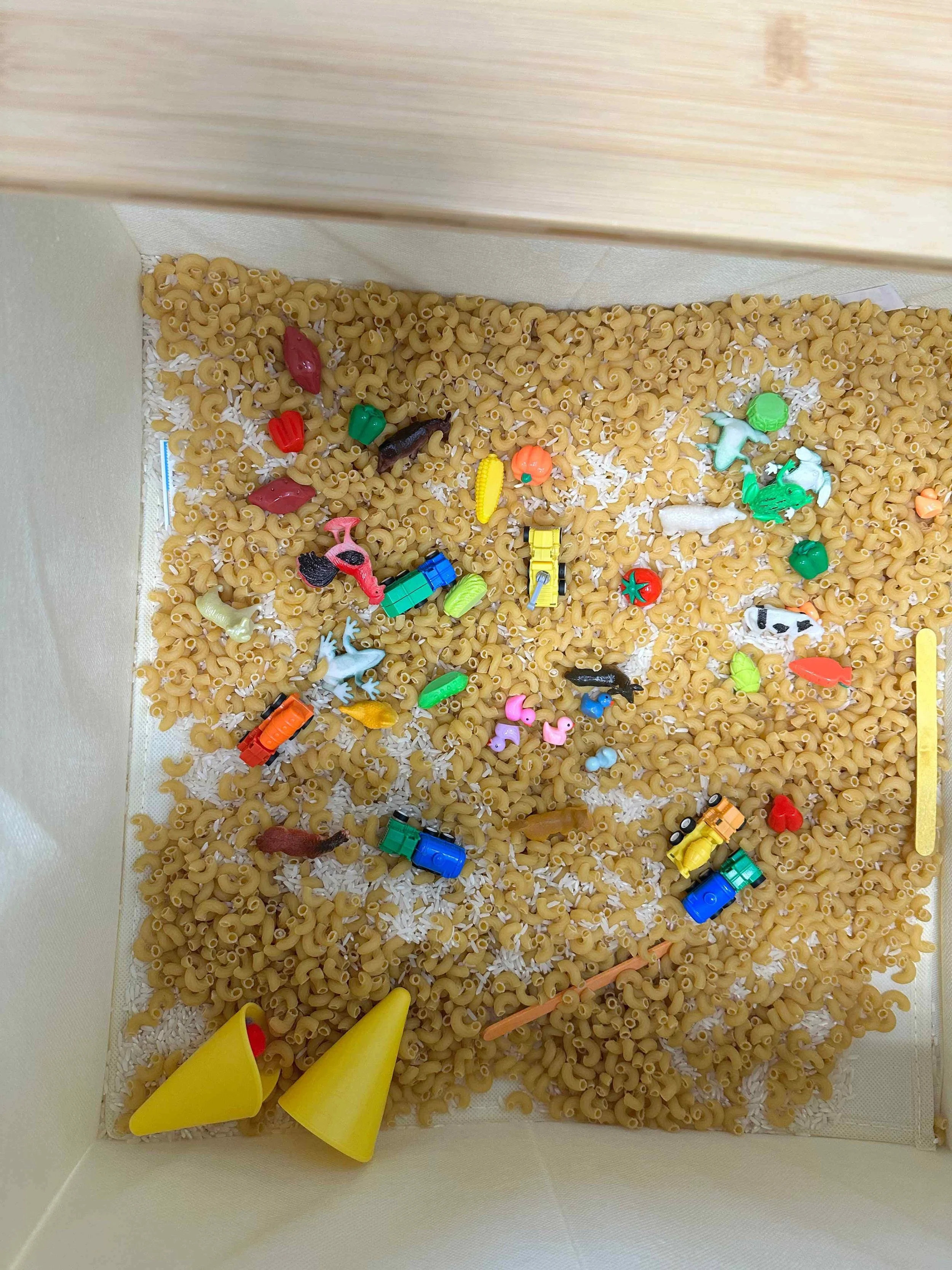 A collection of small plastic toy animals and vehicles scattered on a bed of dry pasta in a white container.