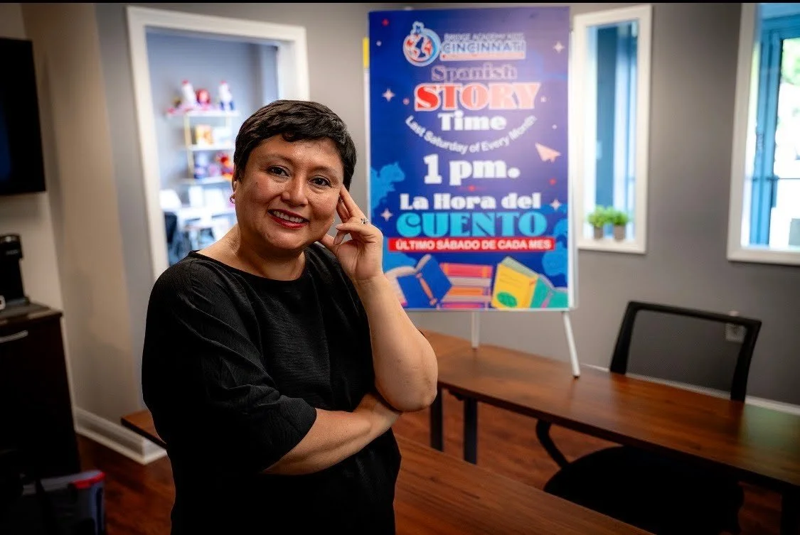 A woman with short dark hair and a black top smiling and posing with one hand on her face in a room with a colorful sign in the background.