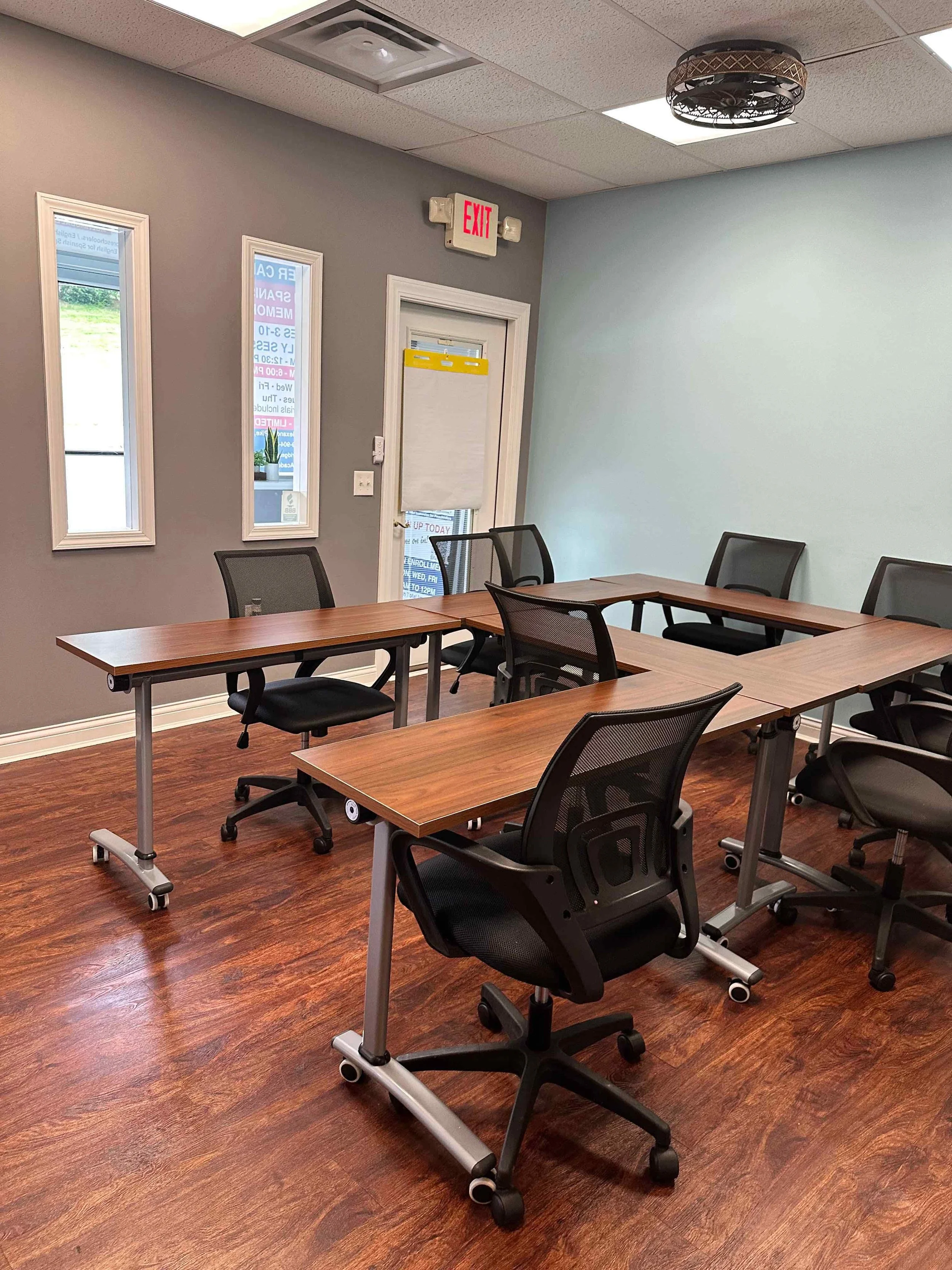 A meeting room with wood flooring, a gray and light blue wall, black chairs with mesh backs, and tables arranged in a U-shape. Two narrow rectangular windows and a door with a window are on the gray wall, with an exit sign above the door. The ceiling