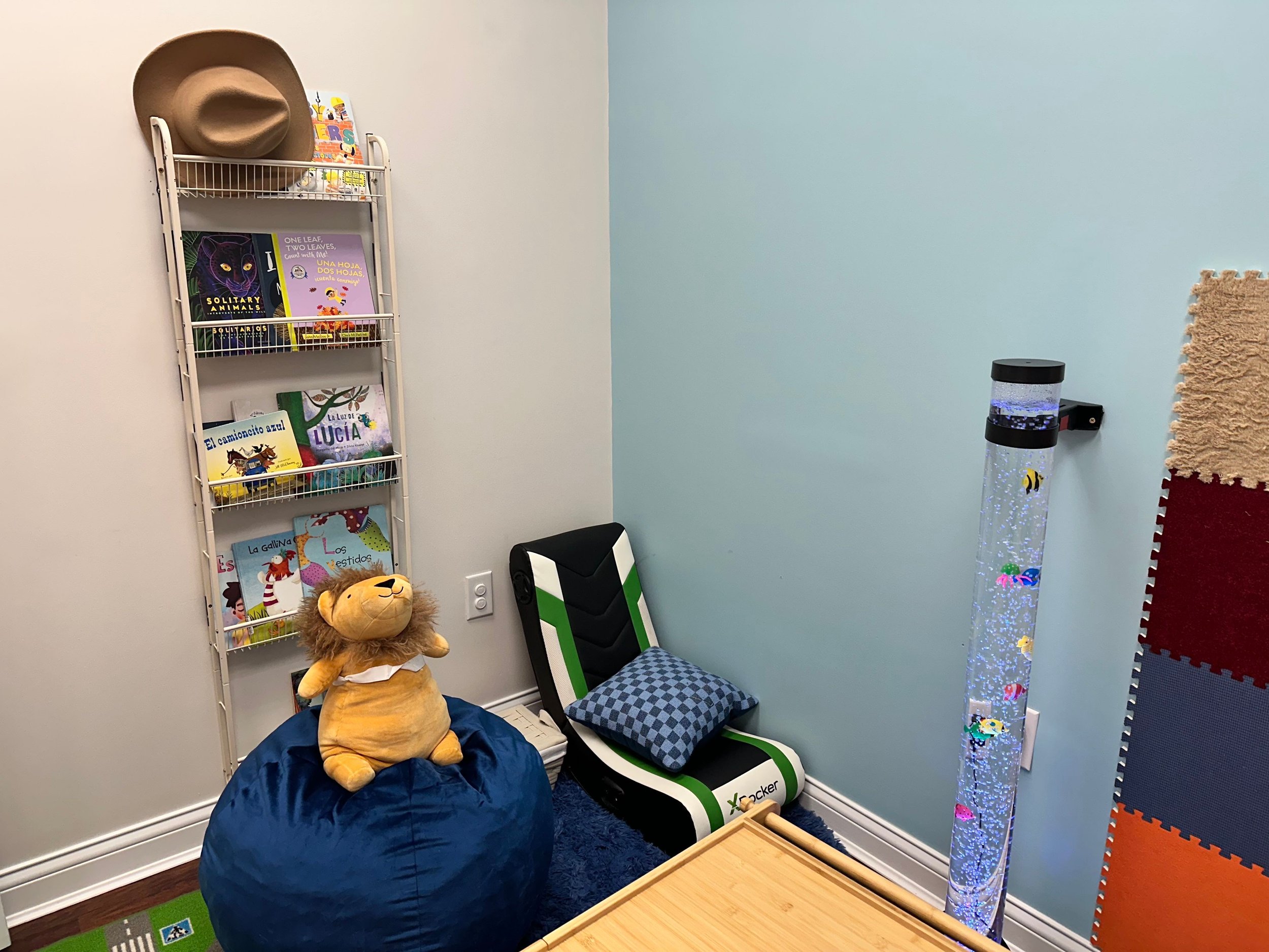 A cozy children's reading nook with a white wire bookshelf holding colorful children's books, a large lion plush toy on a blue bean bag, a small black and white lounge chair with a checkered pillow, a tall bubble tube with colorful fish in water, a b
