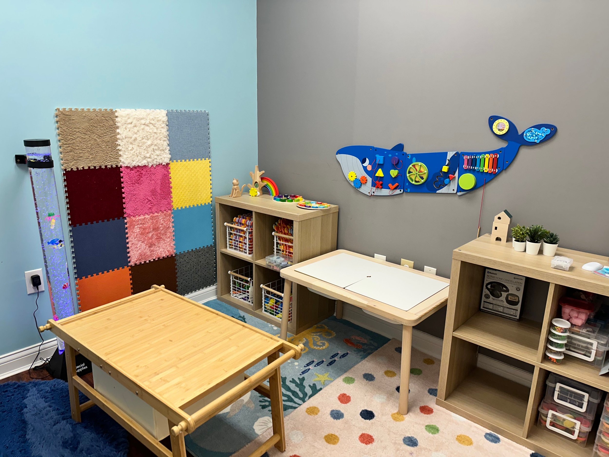 A children's playroom with colorful decor and educational toys. There are two wooden tables, one large and one small, and a storage unit with bins filled with toys. The wall on the right has a large blue whale-shaped activity board with shapes and ob