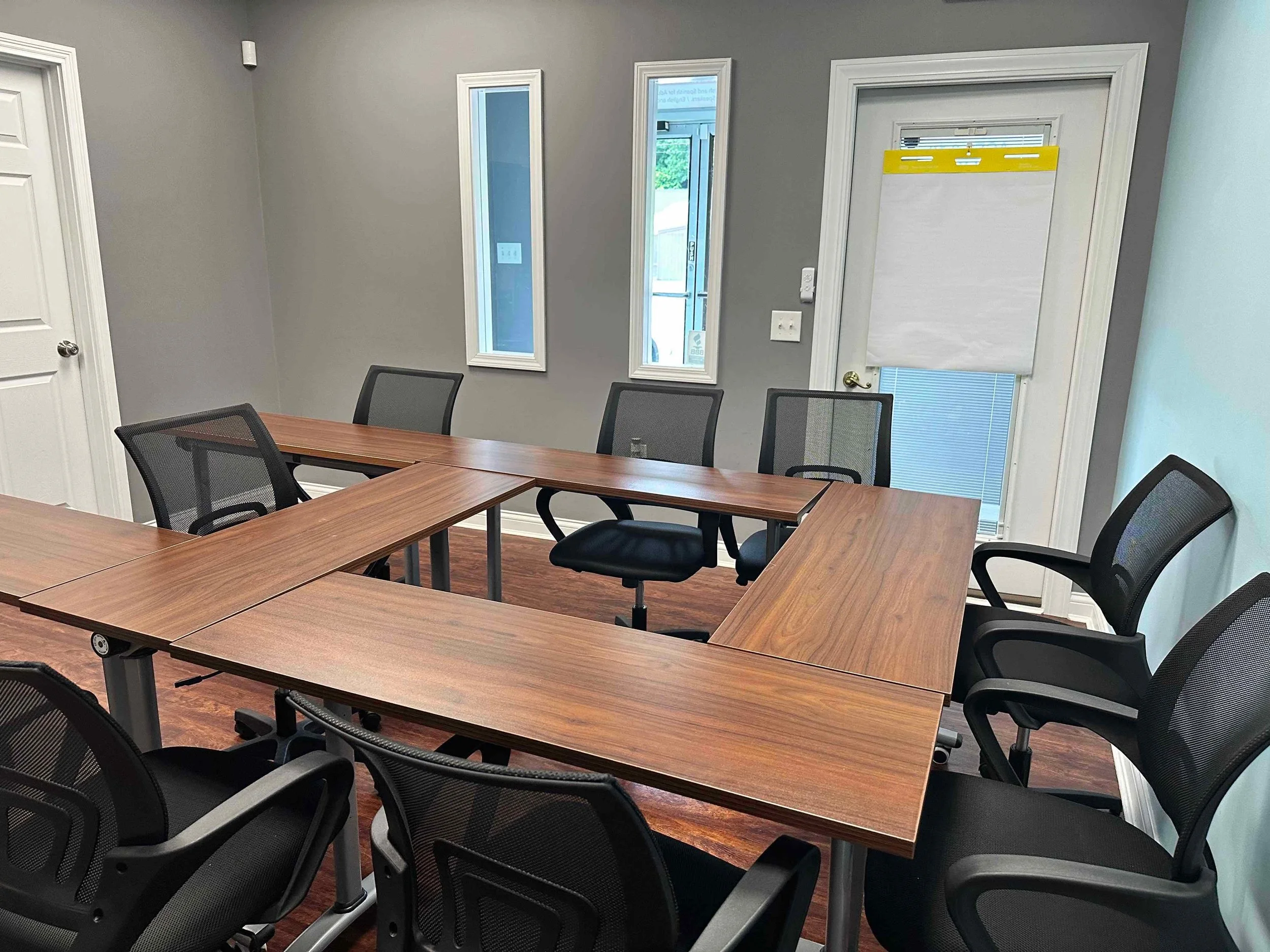 A conference room with a large wooden table surrounded by black mesh office chairs. The room has gray walls, three tall narrow windows, and a door with a white window covering. There is a small white device on the wall and a switch plate.
