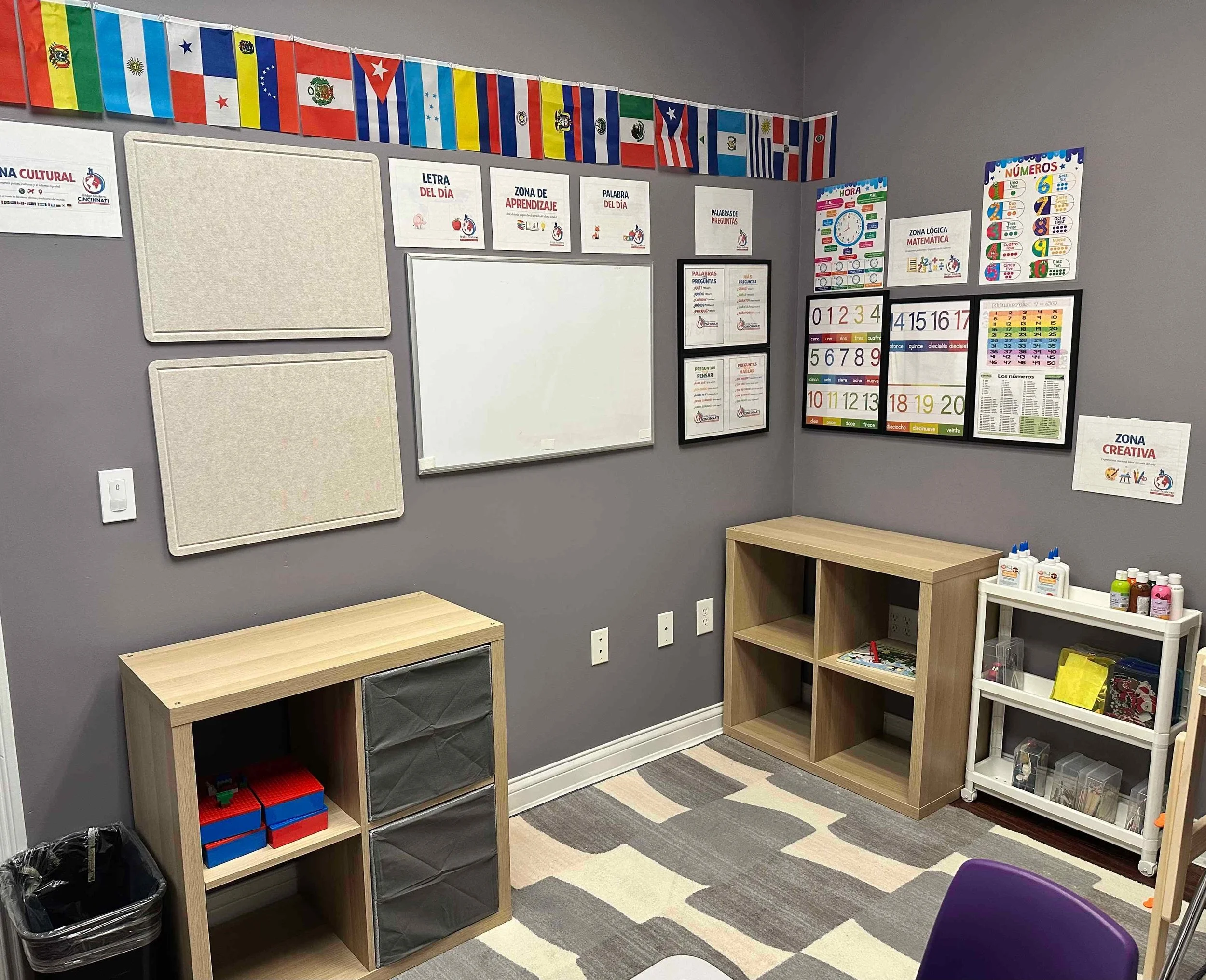 A classroom corner with grey walls and educational posters about numbers, math, and questions. Flags from various countries line the top of the wall. There are empty wooden and plastic shelving units, a colorful striped rug on the floor, and bottles 
