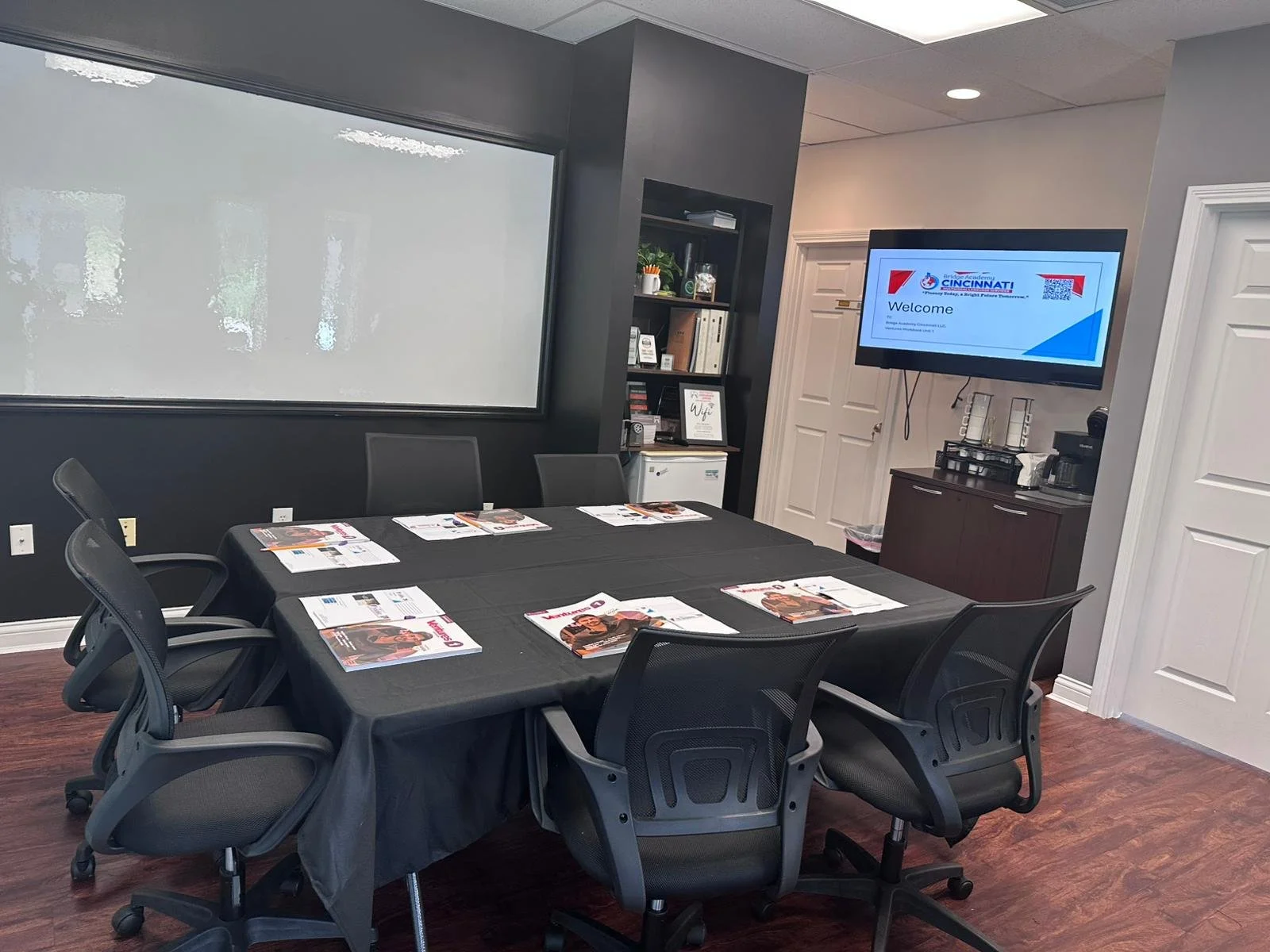 A conference room with six black chairs around a rectangular table covered with a black tablecloth and magazines. There is a large frosted window on the dark gray wall, a bookshelf with decorative items and books, a mini fridge, and a wall-mounted TV