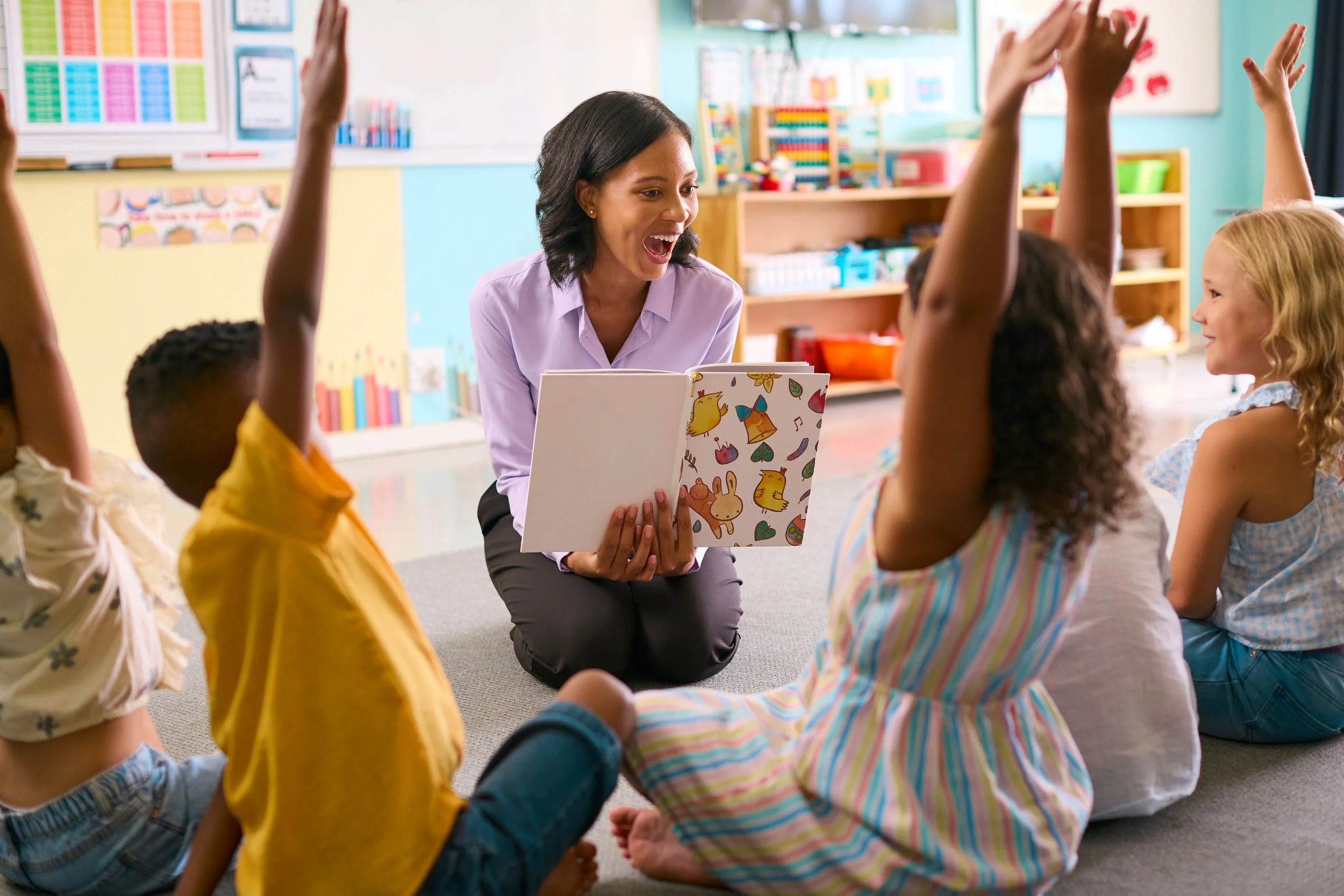 A teacher reading a storybook to a group of young children in a classroom, with some children raising their hands.