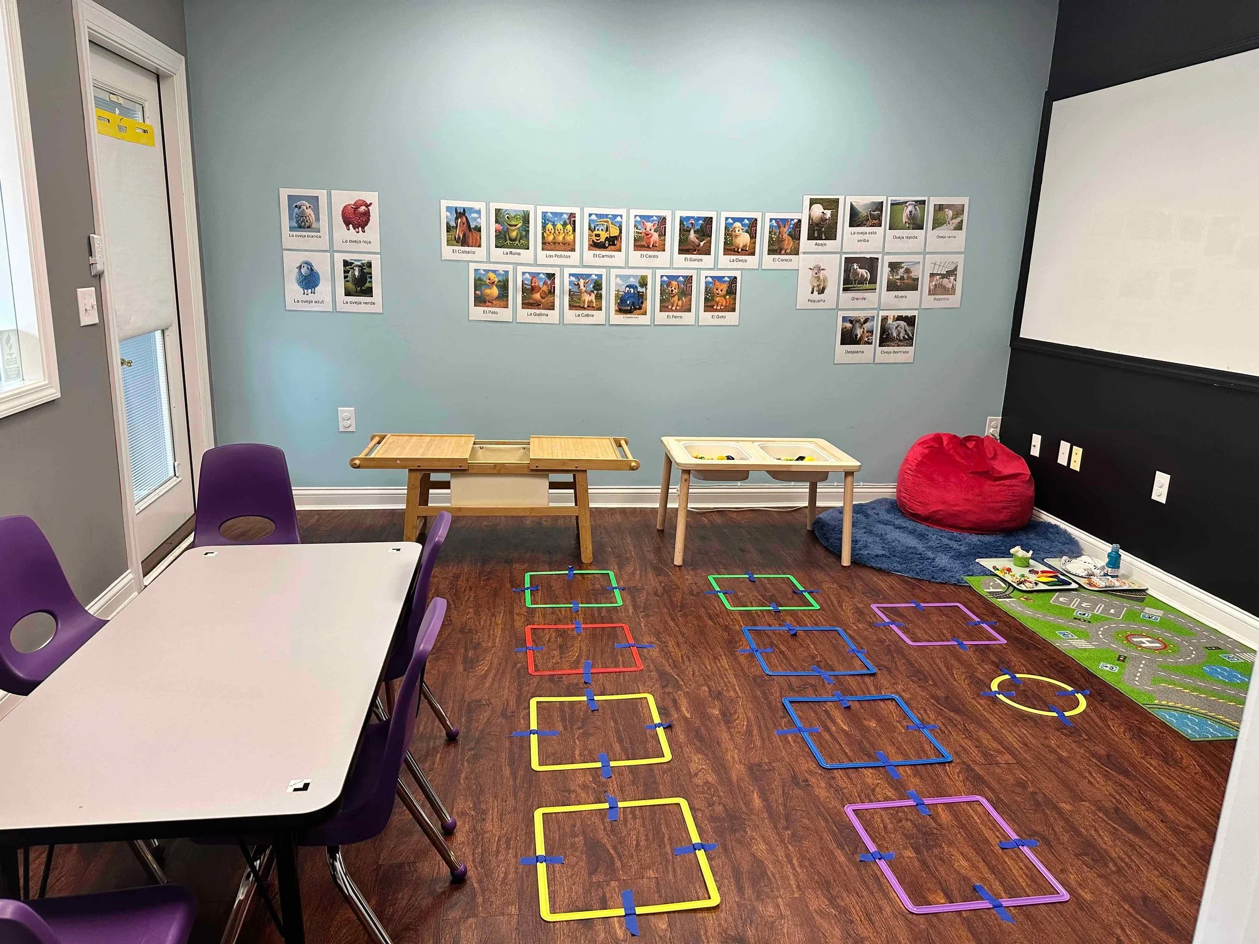 Classroom with colorful hopscotch squares on the floor, purple chairs around a white table, a wall decorated with pictures of animals with their names in Spanish, and furniture including a red beanbag and small tables.