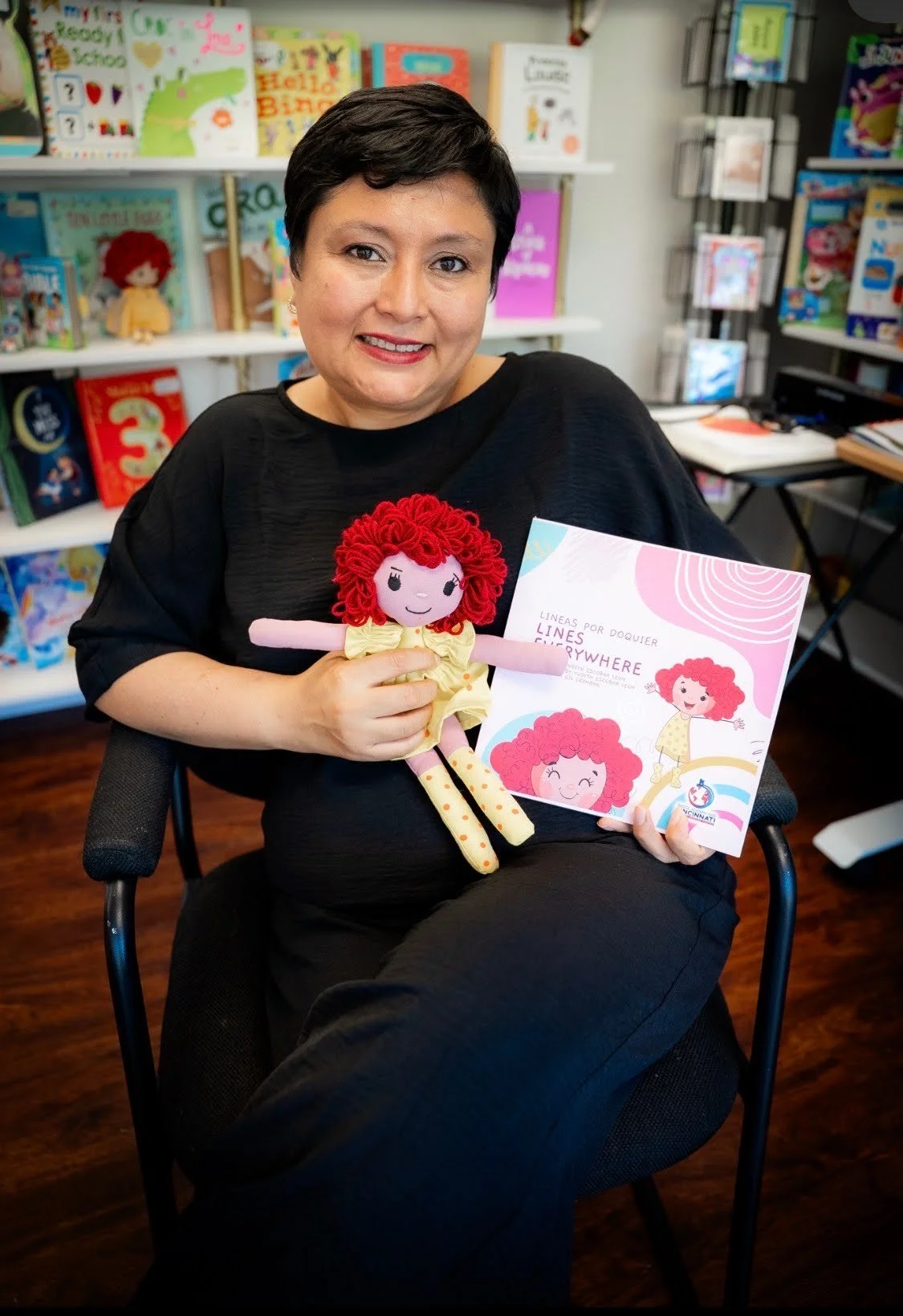A woman with short black hair and wearing black clothing sitting in a chair, holding a red-haired doll with a yellow dress. She is also holding a colorful book with illustrations of the doll and text that reads 'Líneas por doquier' and 'Lines Everywh