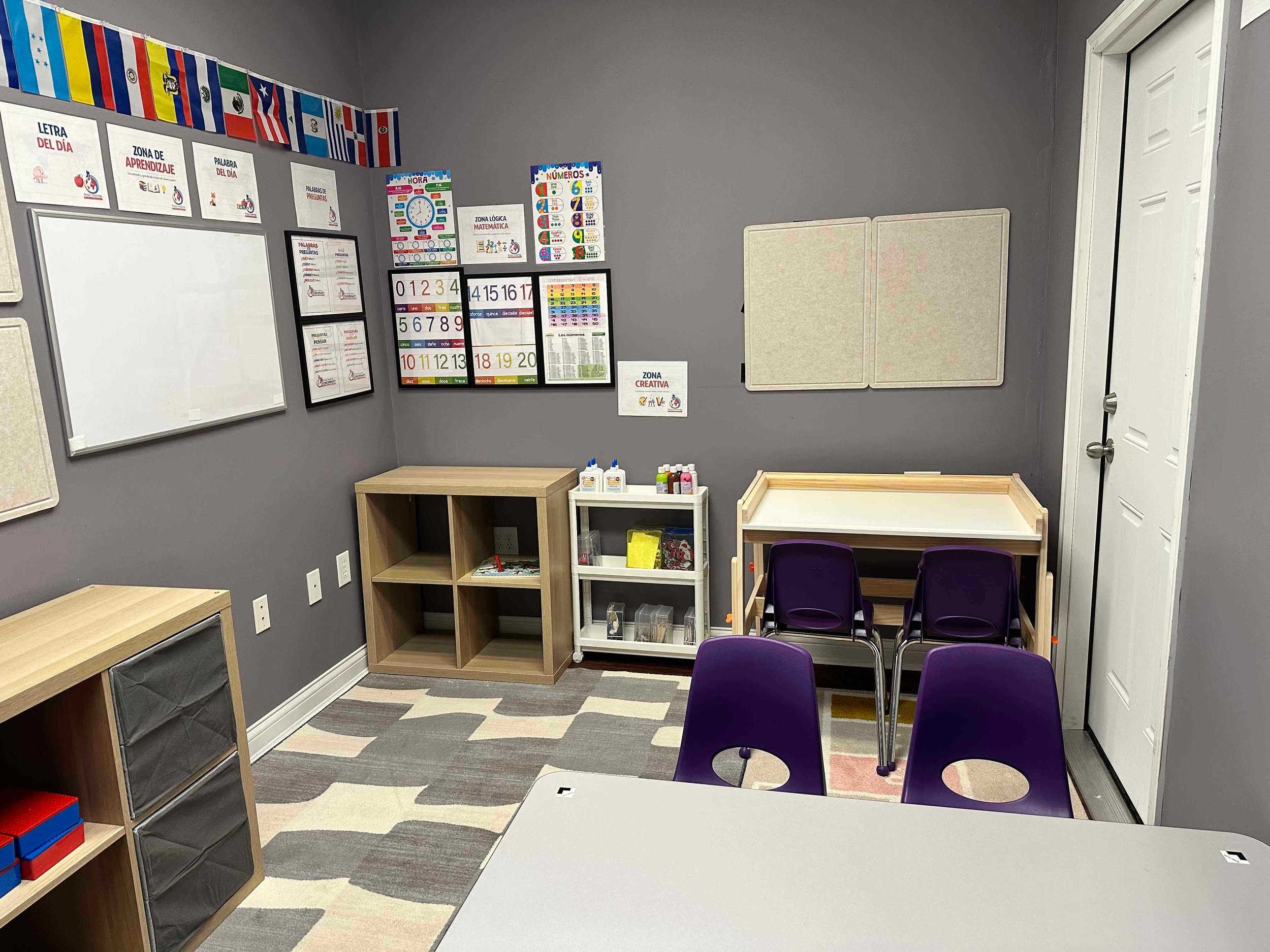 A classroom corner with gray walls, various educational posters on the wall, small wooden and plastic shelves, purple chairs, and a colorful striped rug.