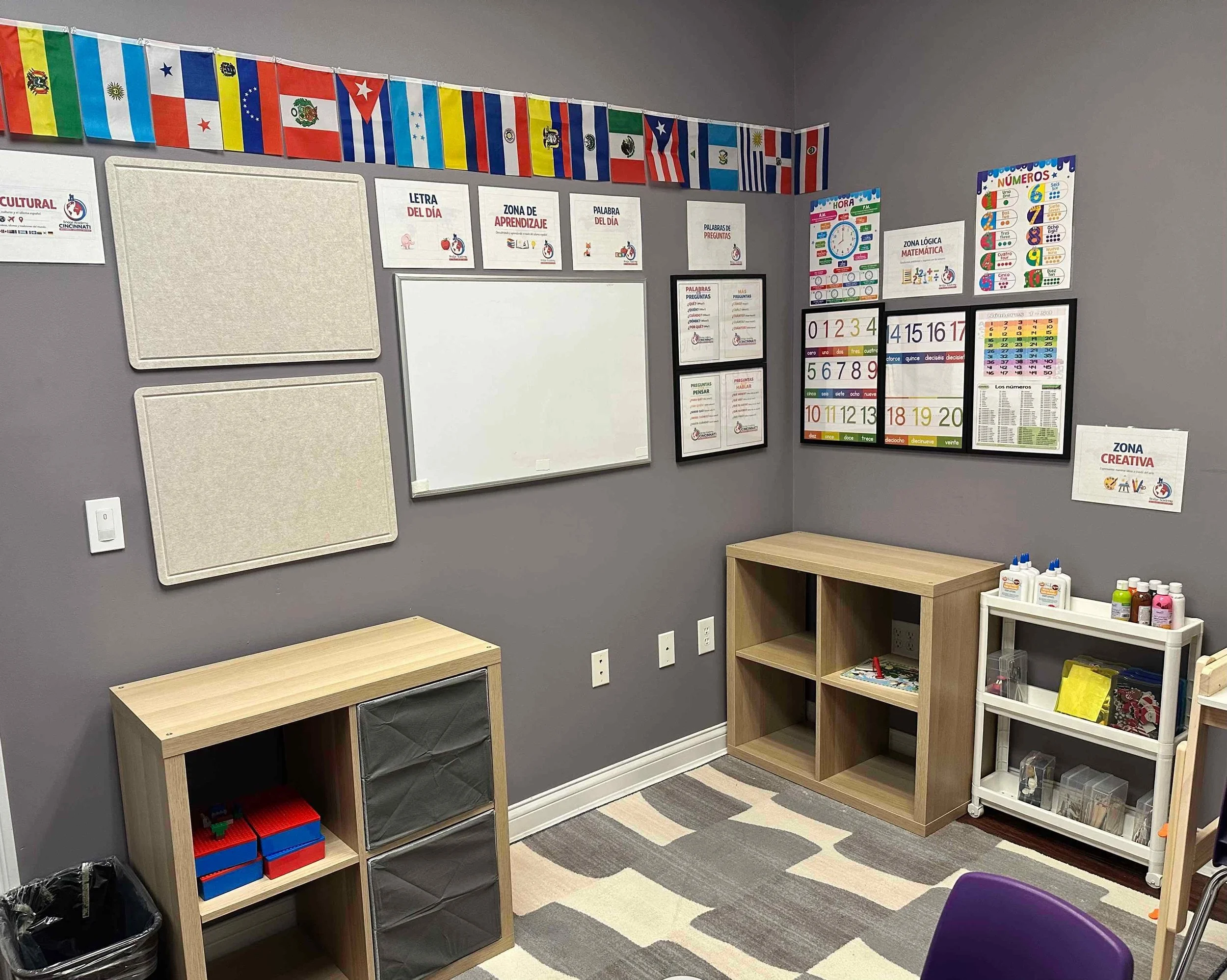 Classroom corner with educational posters on gray walls, wooden shelves, and a cloth-covered table. Colorful flags hang along the ceiling.