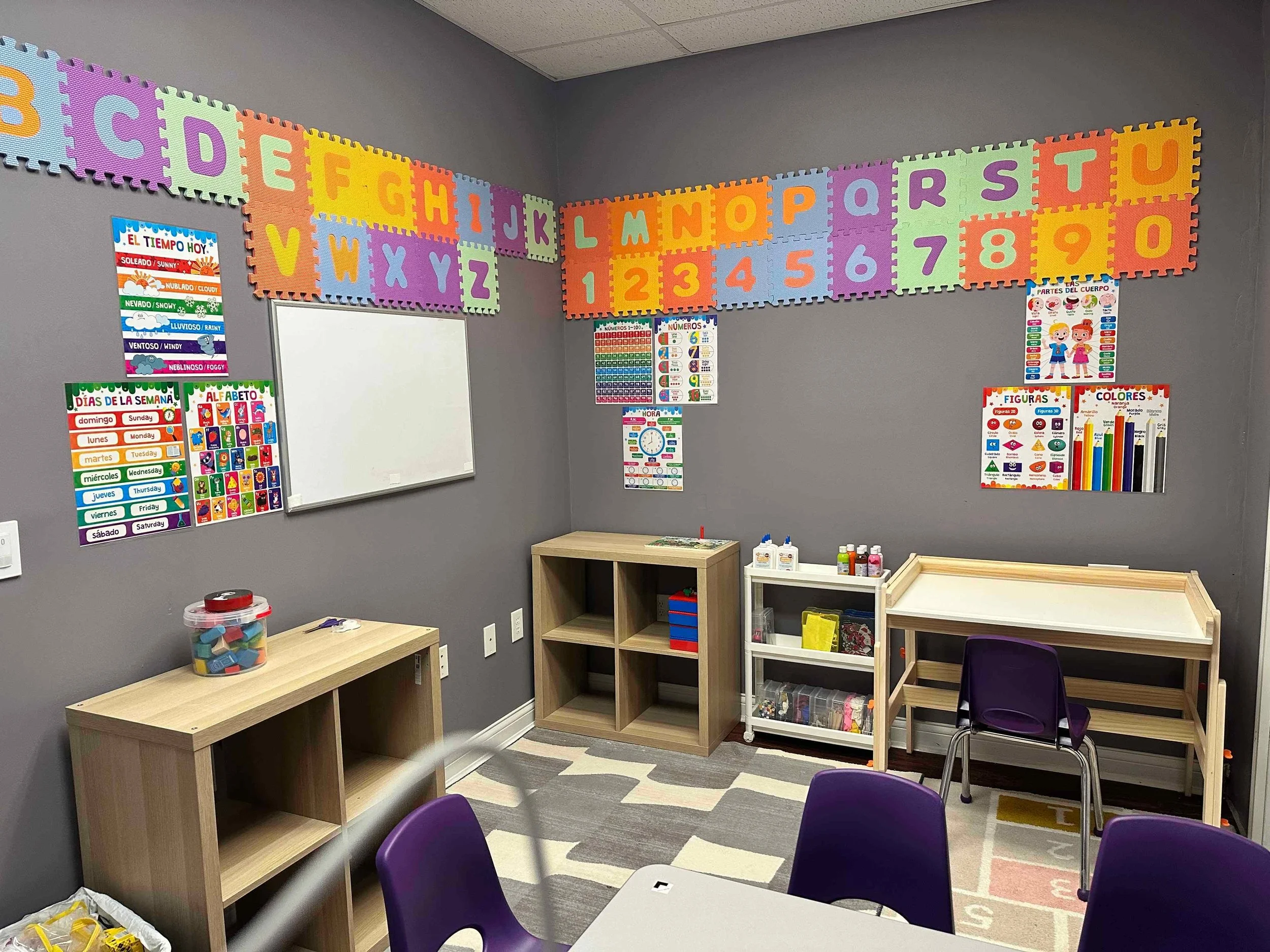 An empty classroom corner with gray walls, decorated with educational posters including the alphabet, days of the week, numbers, parts of the body, figures, colors, and weather. There are several small wooden and white storage shelves, purple chairs,