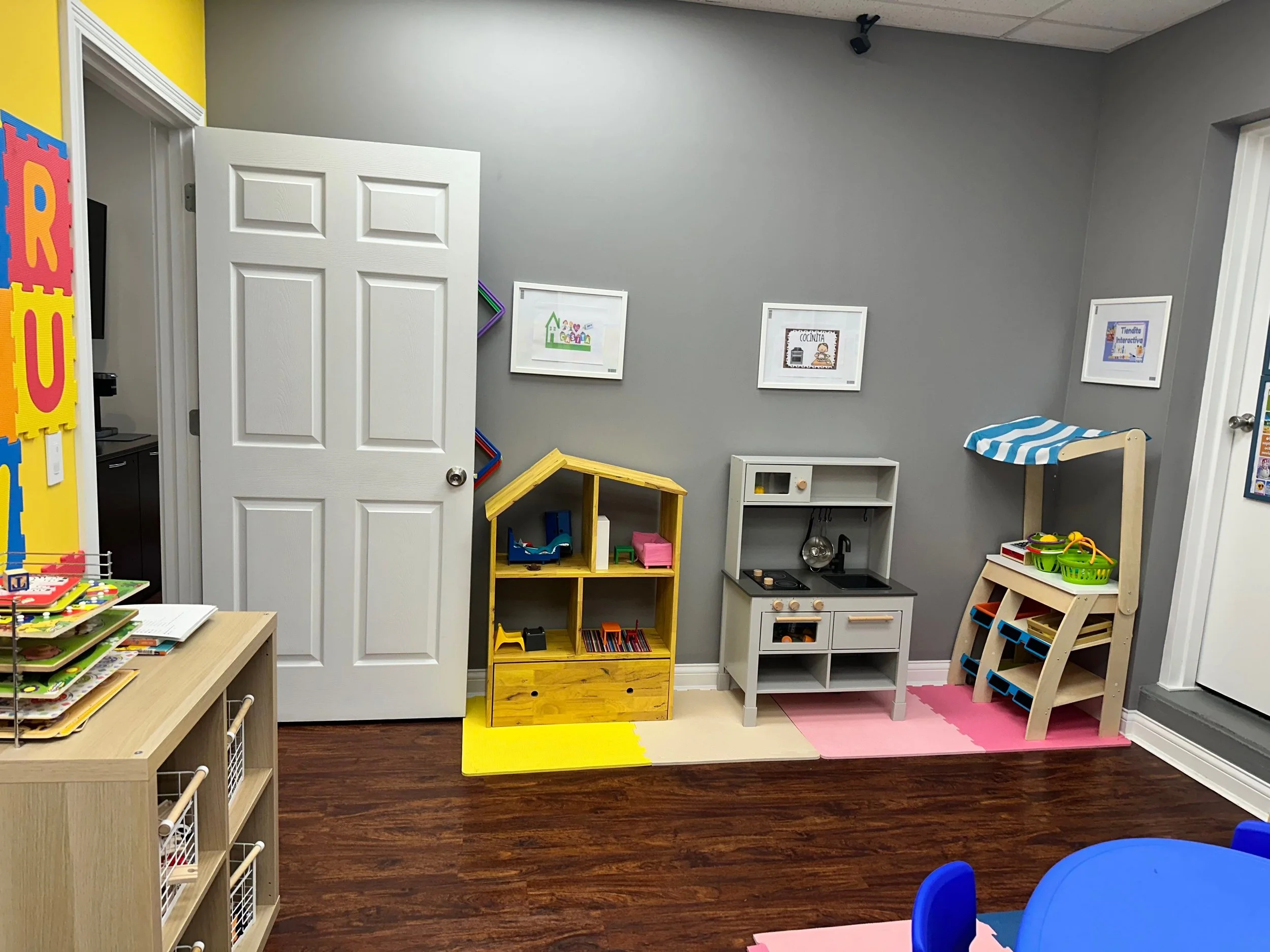 A colorful playroom with a gray wall decorated with framed pictures, a small kitchen play set, and various toys and furniture arranged on a dark hardwood floor.