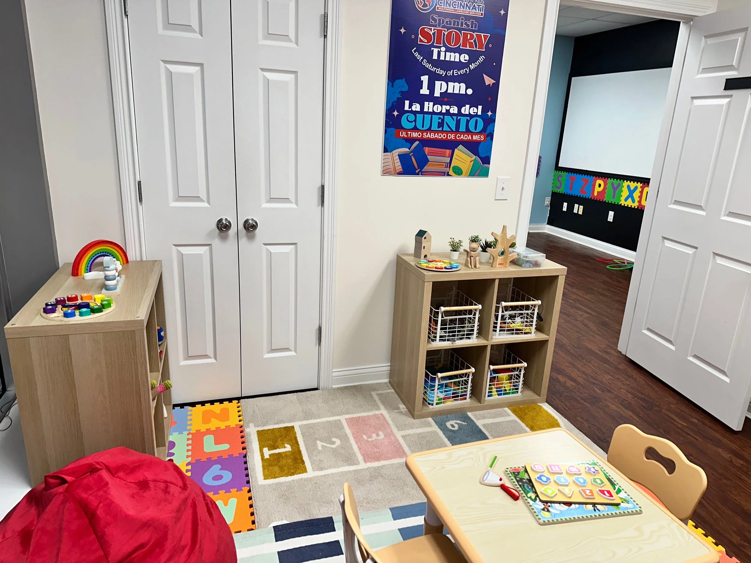 A colorful classroom with a small table and chairs, a rainbow and stacking toy on a side table, a children's puzzle rug, and educational posters on the walls.