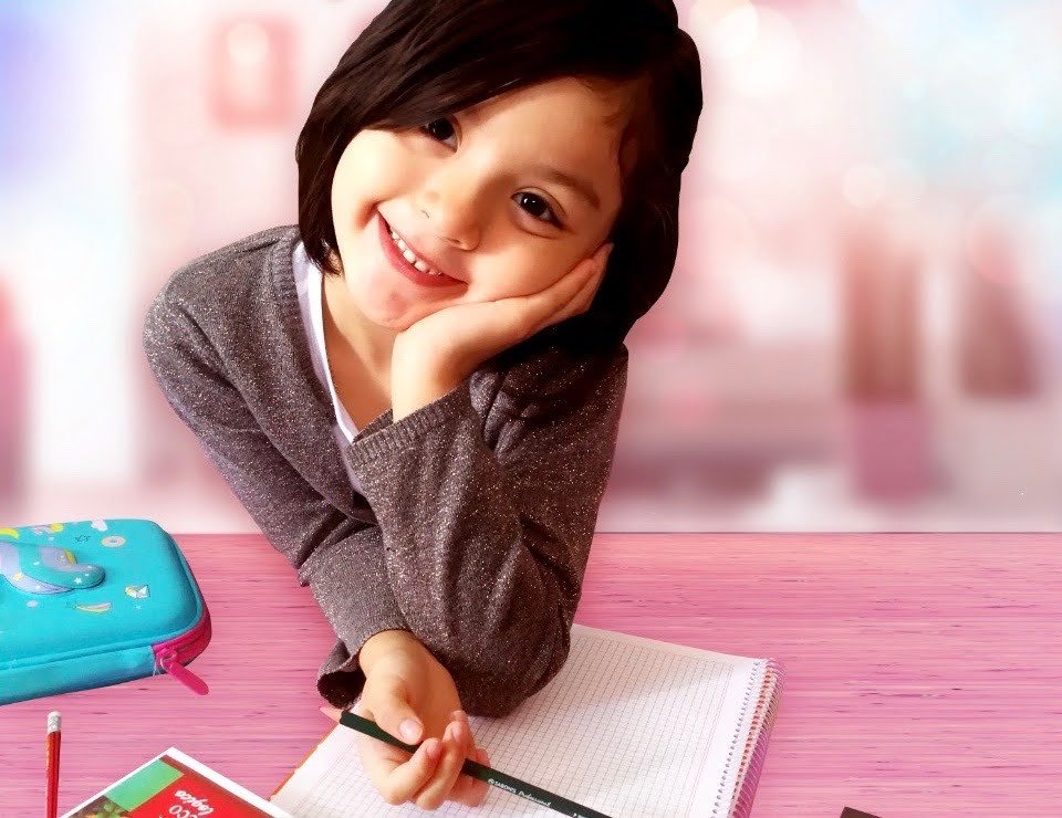 Smiling young girl with dark hair resting her face on hand at a pink desk, with a blue pencil case, notebook, and pencil in front, in a blurred pink background.