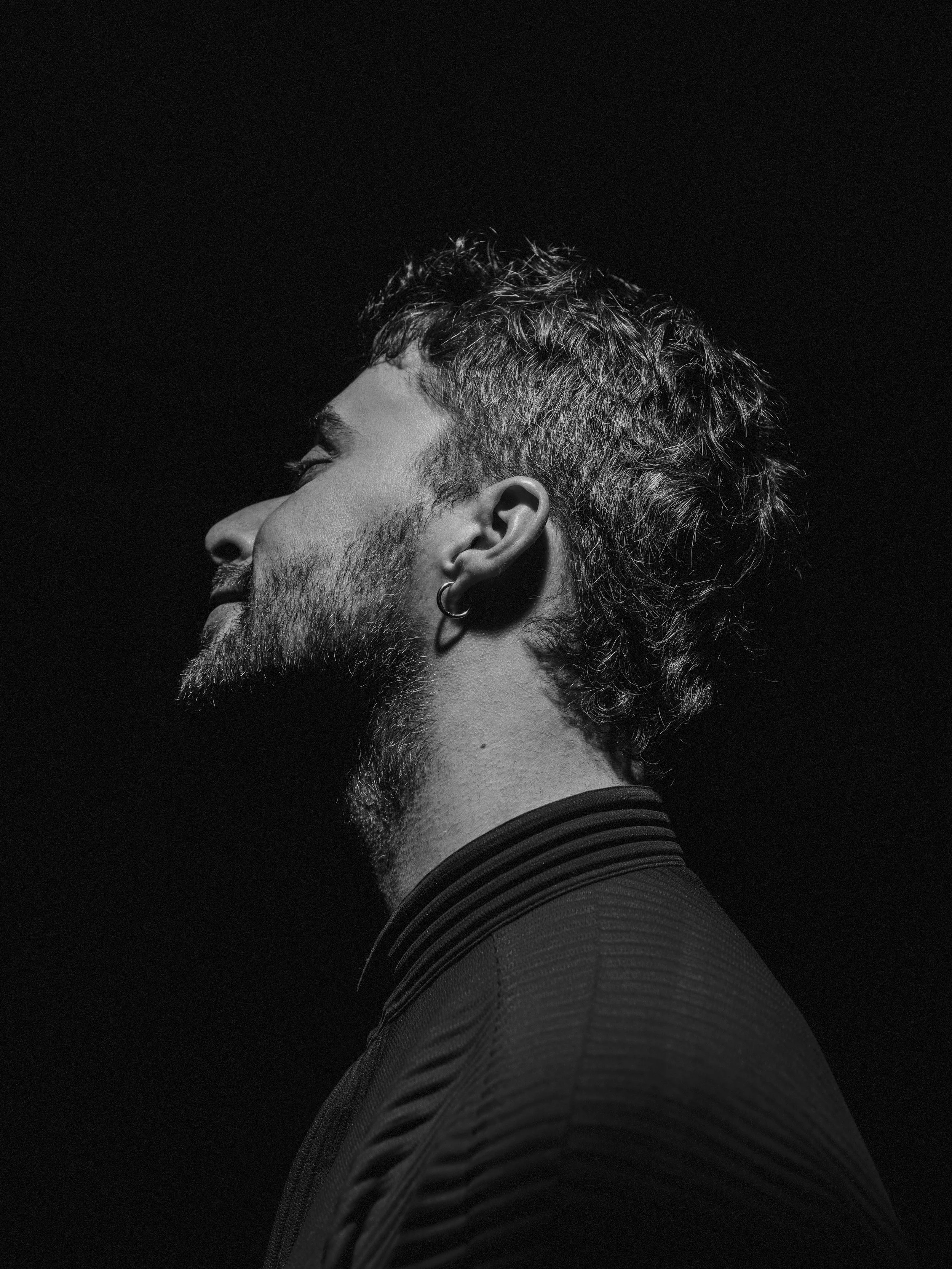 Profile of a man with a beard and hoop earring, wearing a striped shirt, against a black background.