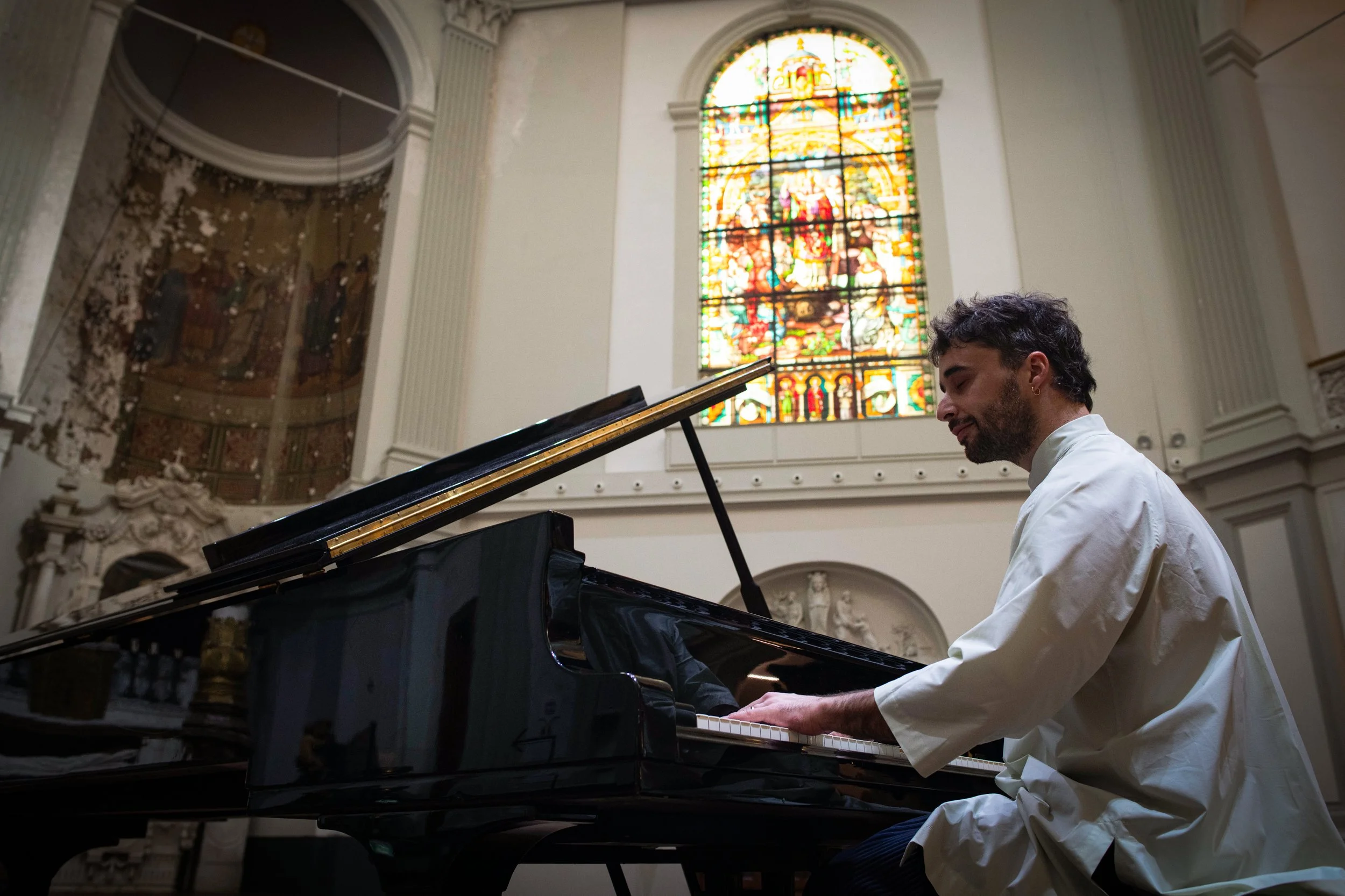 A man playing a grand piano in a church with stained glass windows featuring religious artwork and figures.
