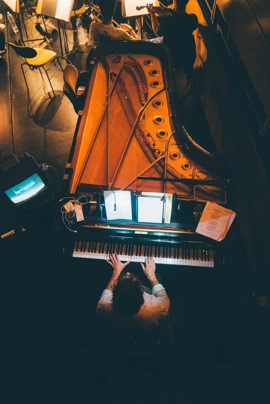 Raúl da Costa playing a grand piano on a stage, with sheet music and digital screens, while an audience and chairs are visible in the background.