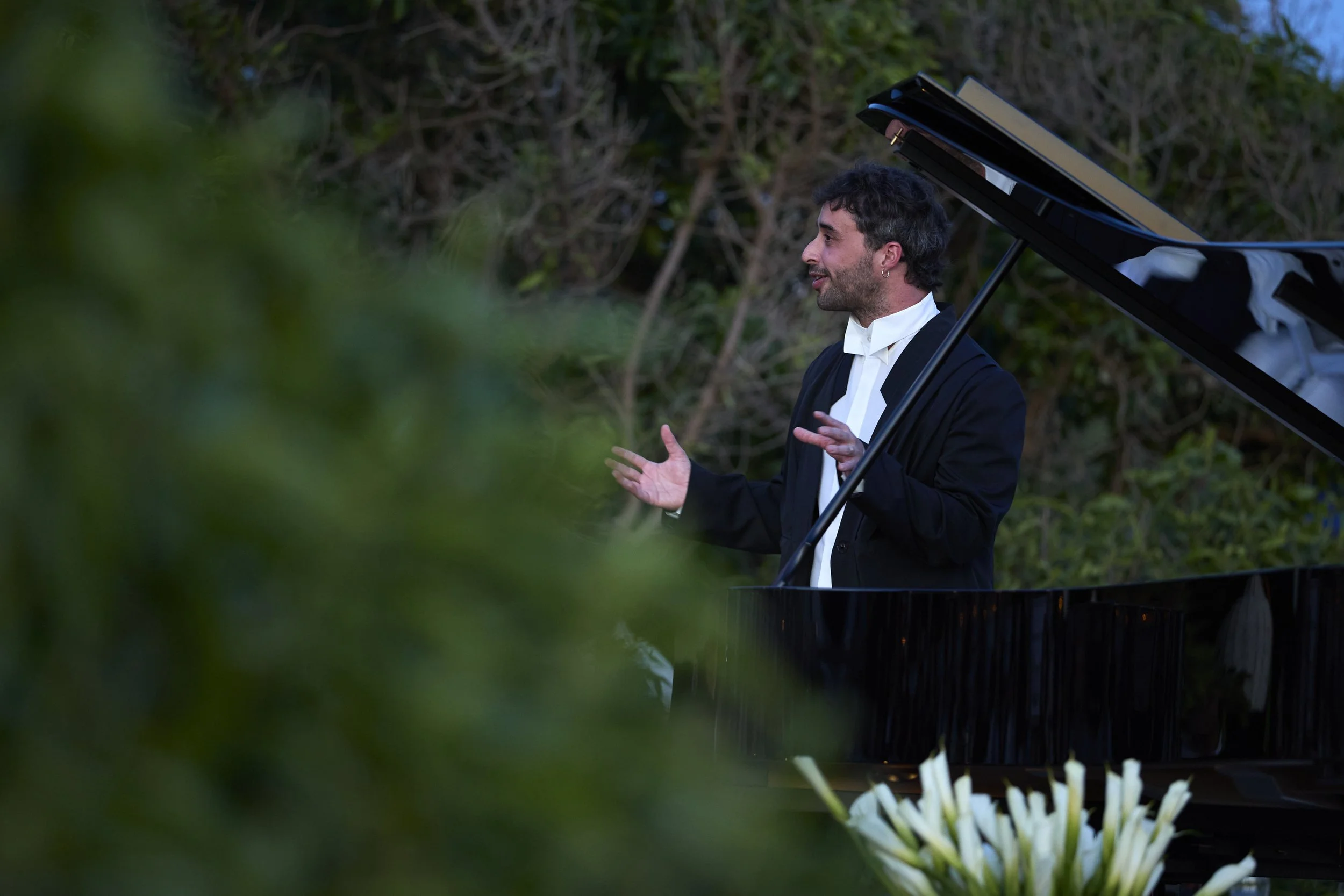 A man with dark hair and a beard, wearing a black suit and white shirt, stands next to a black grand piano outdoors surrounded by greenery. He appears to be speaking or explaining something.