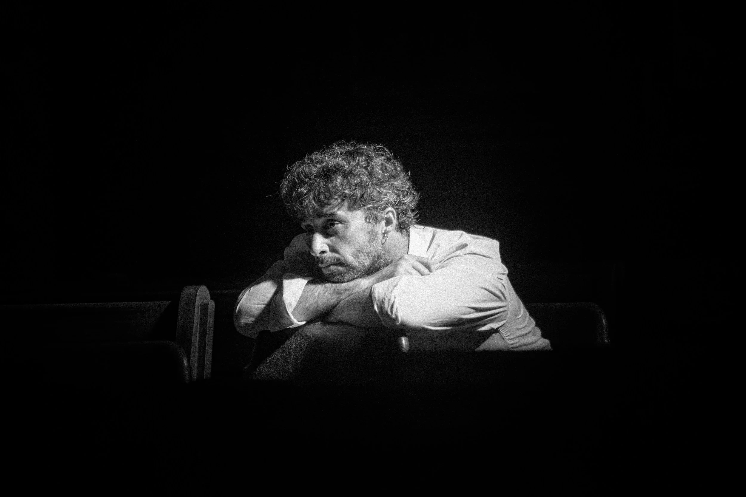 A black and white photo of a man with curly hair and a beard resting his arms and head on a table, looking down.