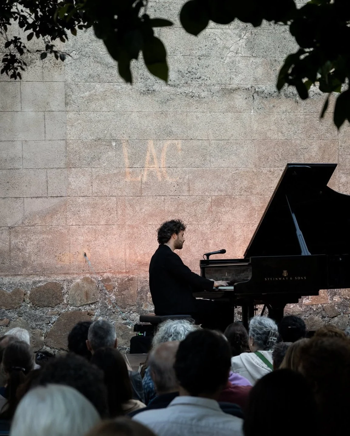 A man playing a grand piano during a concert outdoors with an audience watching, behind a stone wall with the letters 'MC' projected on it.