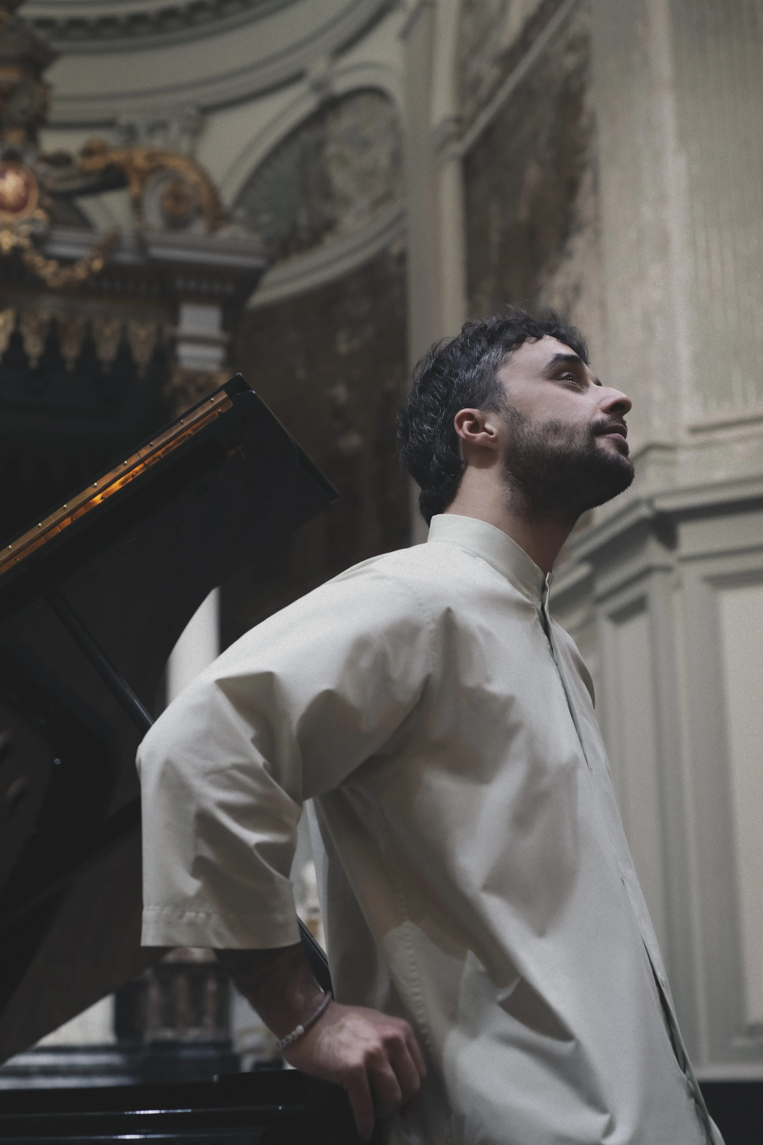 A man with dark curly hair and a beard wearing a white shirt, standing in a room with ornate ceiling details, near a grand piano.