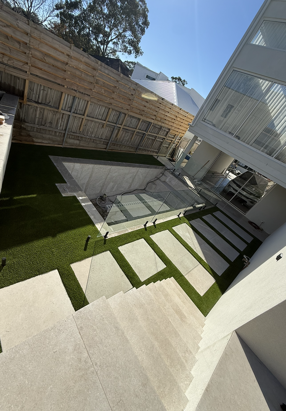A backyard with a glass fence, green artificial grass, and a swimming pool under construction. There are stairs leading down from the house, a wooden fence, and a modern white building with large windows.