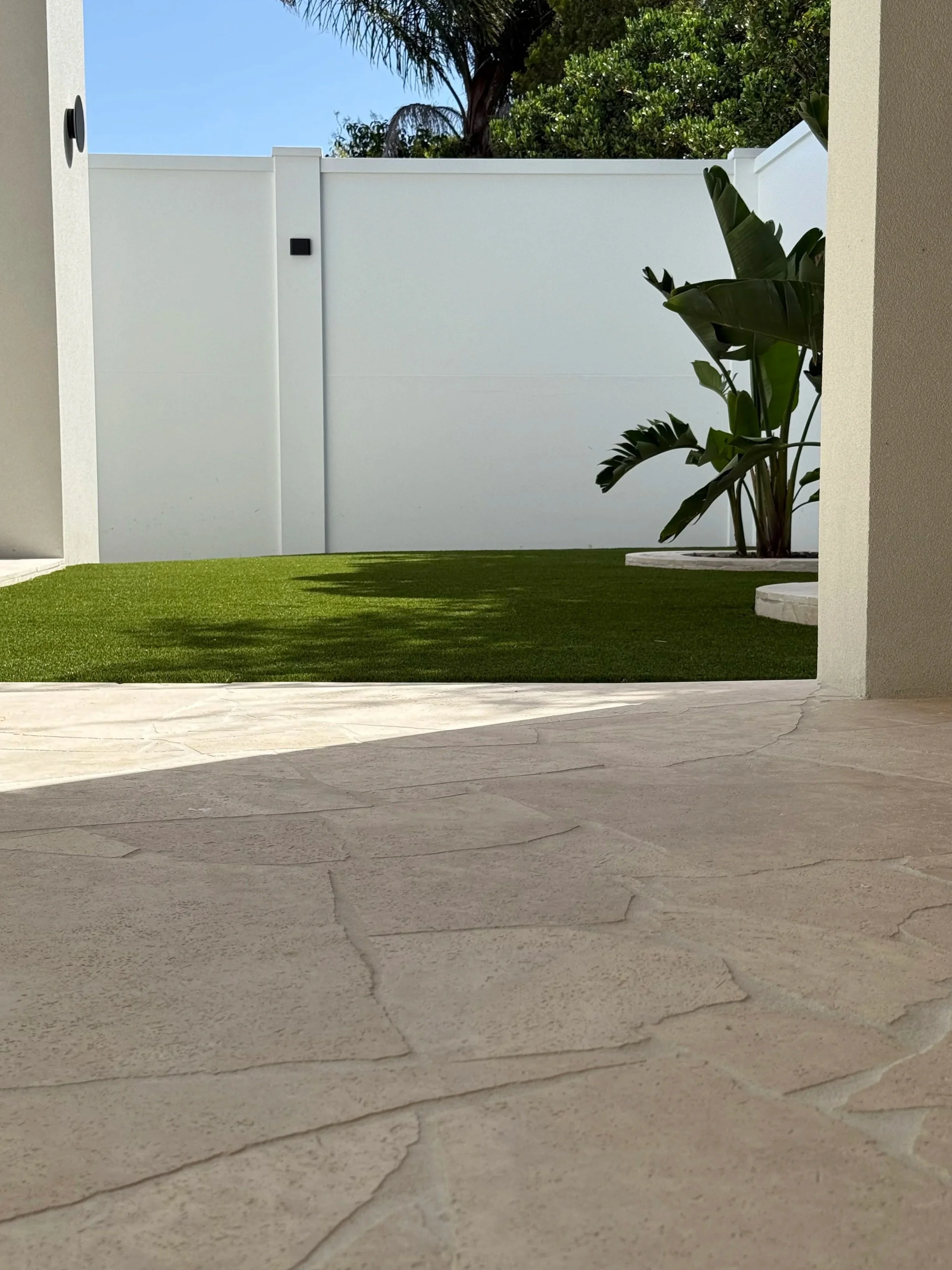 A backyard with beige stone patio flooring, green artificial grass, a white privacy fence, and tropical plants including banana plants and palm trees, under a clear blue sky.