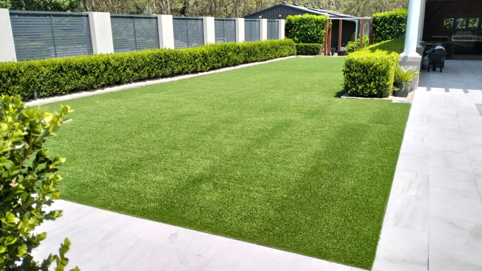 A backyard with a well-maintained artificial grass lawn, bordered by neatly trimmed shrubs and a gray fence. There is a patio area with chairs and a small garden shed in the background.