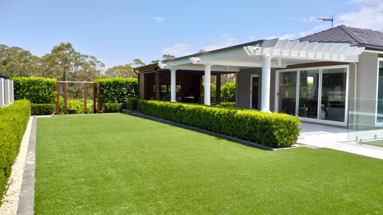Modern backyard with neatly trimmed bushes, a green lawn, and a house with a covered patio area and large glass sliding doors.