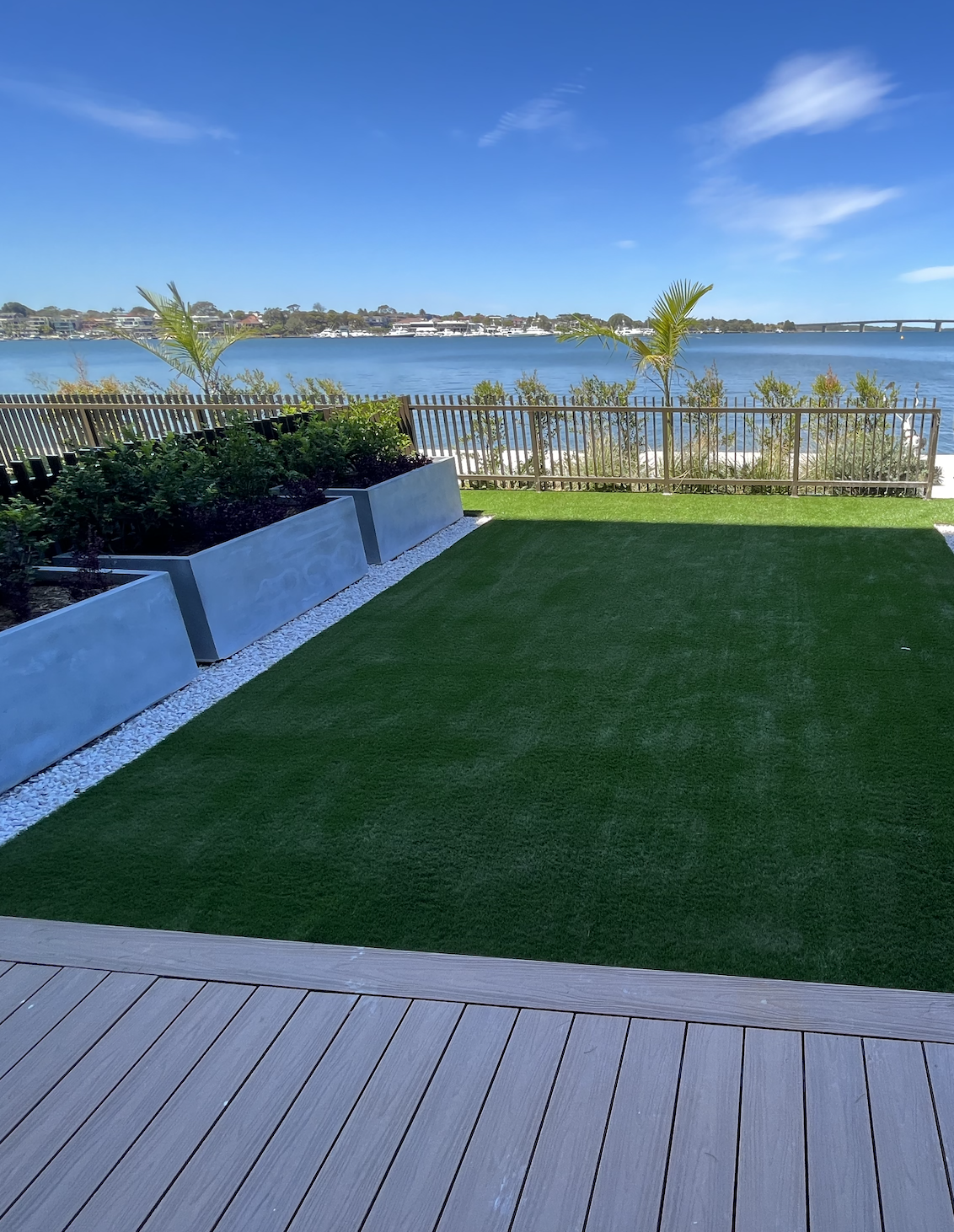 Backyard with artificial grass, potted plants, and a wooden fence overlooking a body of water with a bridge in the distance and a blue sky with few clouds.