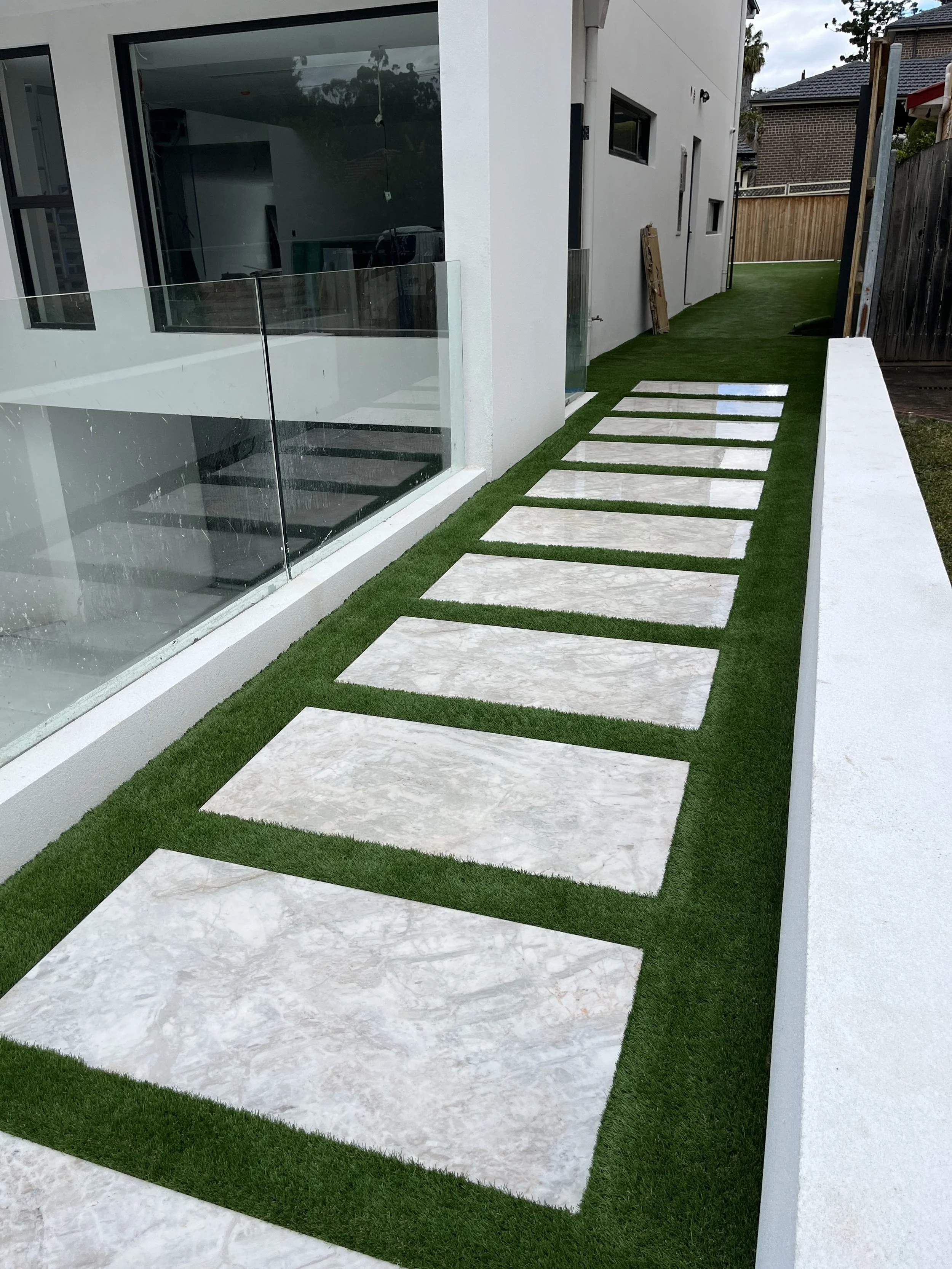 Modern house exterior with a white wall, large window, glass railing, and a pathway with rectangular stone slabs set in artificial grass