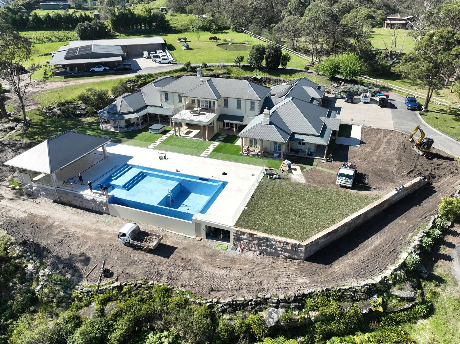 Aerial view of a large house under construction with a swimming pool in the backyard, construction workers, and construction vehicles. The house features multiple gabled roofs, large windows, and a patio area with a lawn and a pond nearby.